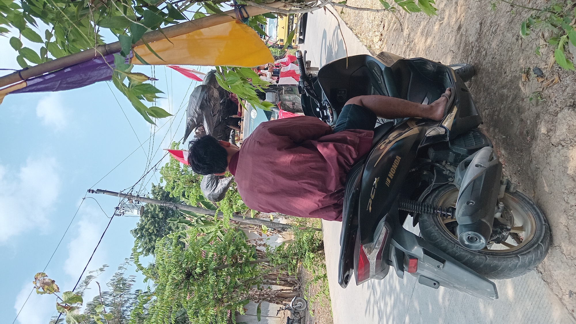 In Belor, Indonesia, a person sits on a black Honda PCX scooter on a dirt and asphalt road. The individual is wearing a maroon bomber jacket and dark shorts, with one foot in a flip-flop resting on the scooter's footboard. The scooter has a black finish with gold accents and "PCX" and "HONDA" branding. Behind the scooter, a truck is visible, laden with what appears to be trash bags, some adorned with red and white Indonesian flags. Other vehicles, including a yellow truck, are on the road. The environment is rural, with lush green trees, banana trees, and vegetation lining the road. Electrical wires and poles are prominent in the background, and a transformer is mounted on one pole. The sky is bright blue with scattered clouds, suggesting a sunny day.