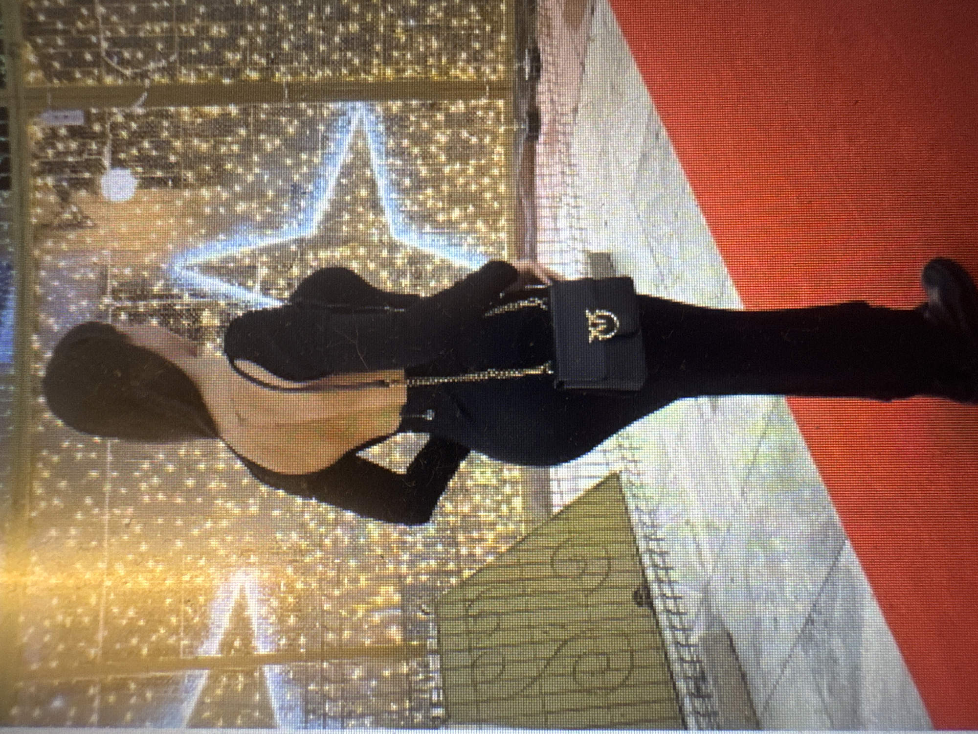 The image shows a woman standing on a red carpet in front of a backdrop of numerous small, yellow lights and a large blue star outline made of lights. The woman is wearing a black, long-sleeved dress with a very low back, exposing her upper back. She is carrying a small black handbag with a gold chain strap and a distinctive gold buckle with a circular emblem. She has long, dark hair that is styled down her back. The background appears to be an outdoor setting, possibly a plaza or street, adorned for a festive occasion with bright lighting. There is a large, yellow triangular structure with a swirled pattern to the left of the woman. The overall impression is of an event or celebration. The question asks to include the city of Murcia, Spain, however, there are no visual cues within the image that specifically identify the location as Murcia.