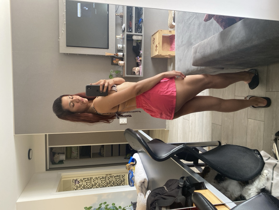 A woman with long, reddish-brown hair, wearing a patterned bikini top and bright pink shorts, is captured taking a full-body mirror selfie. She holds a dark smartphone in front of her face, with her eyes closed or looking down, and her lips slightly puckered in a pose. She is also wearing black flip-flops.
The setting appears to be a modern indoor room, likely a living area or bedroom in Buenos Aires, Argentina, featuring light-colored flooring. Reflected in the mirror behind the woman is a grey wall with a large flat-screen TV mounted on it. To the right of the TV, a multi-tiered shelving unit displays various decorative items, including small sculptures and containers. Below these shelves, a small, light-colored wooden pet bed with a pink cushion is visible, along with a section of a grey sofa. In the foreground, a black office chair with a mesh back is partially visible, along with a white towel and a black bag resting on a surface. Some blue and yellow goggles or similar eyewear are also seen near the towel.
The lighting suggests it is daytime. No discernible text is clearly readable on the TV screen or any other objects. The scene conveys a casual, personal moment.