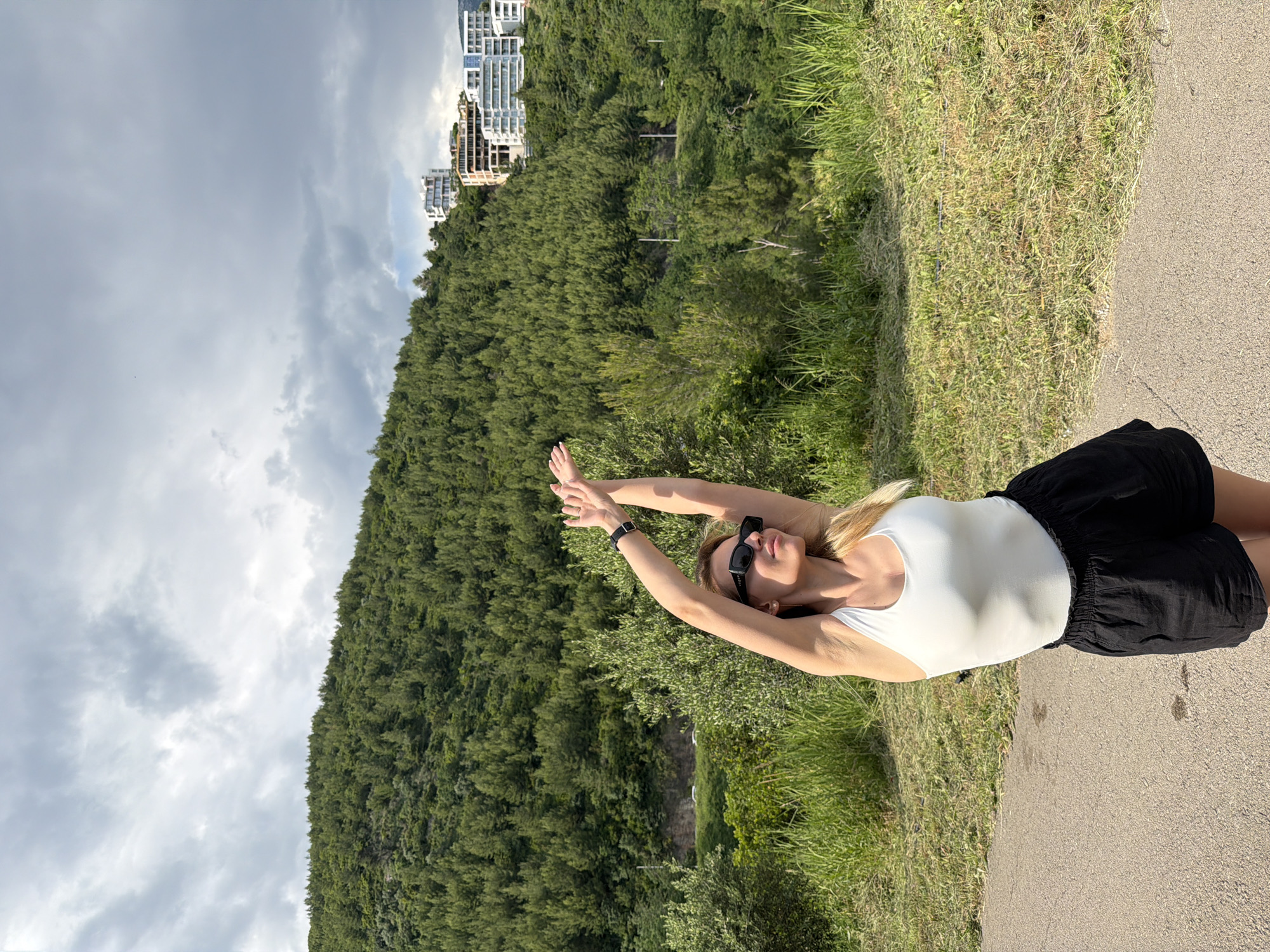 A woman in a white leotard and black shorts stands on a paved path, extending her arms above her head. She wears sunglasses and a wristwatch. Lush green vegetation covers a hillside behind her, with buildings visible at the top. A cloudy sky forms the backdrop. The location is possibly near the city of Budva, Montenegro, given the vegetation and architecture.