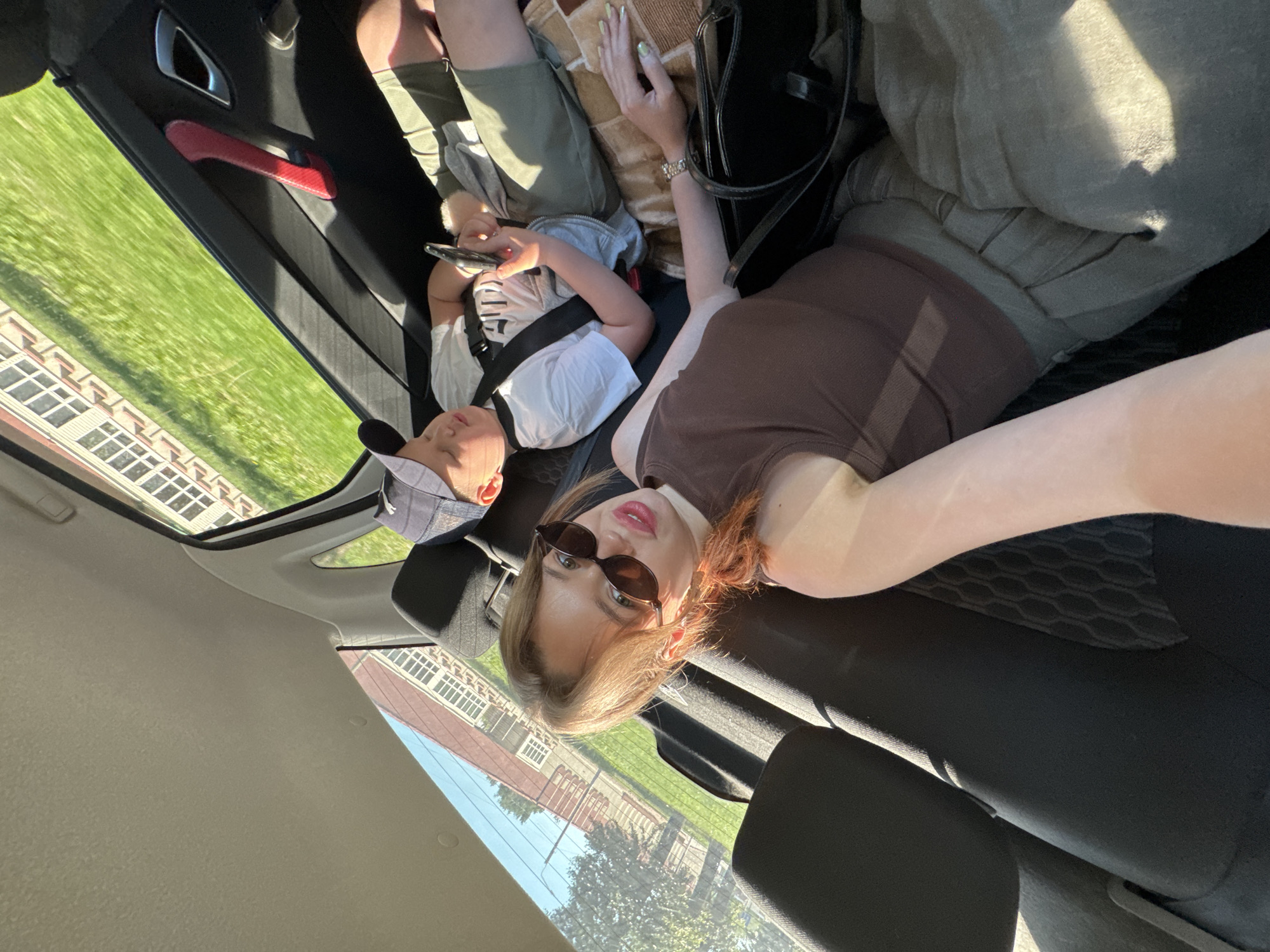 In the backseat of a car, a young woman wearing sunglasses and a brown tank top is taking a selfie. Beside her, a young boy wearing a baseball cap and a white t-shirt is sleeping, holding a mobile phone. They are on a road trip, with a glimpse of a building and green grass visible through the car window, suggesting a suburban or rural setting. The woman's arm and hand, adorned with a watch and mint-green nail polish, are visible in the foreground, along with her black handbag. The car interior appears to be fabric-upholstered seats. The time of day is likely afternoon, given the sunlight. The specific location in Chaikovsky, Russia, is not discernible from the image.