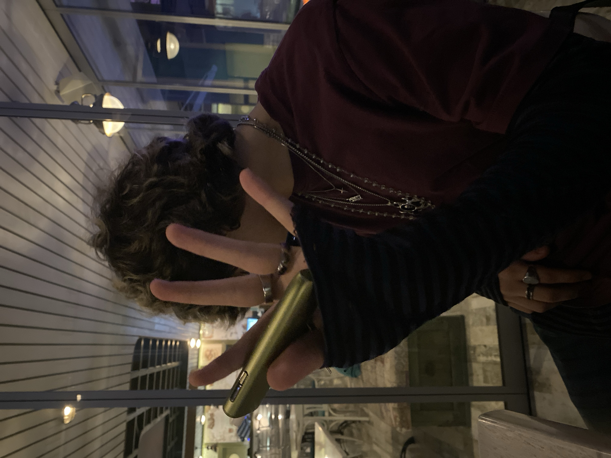 An image shows a person holding a vape pen in front of their face, gesturing with two fingers in a peace sign. The person has curly brown hair and is wearing a dark red shirt with a long-sleeved black undershirt and several silver necklaces. They are also wearing rings on both hands. The background shows a street scene at night with streetlights and buildings, suggesting an outdoor setting. The location is Stara Zagora, Bulgaria.