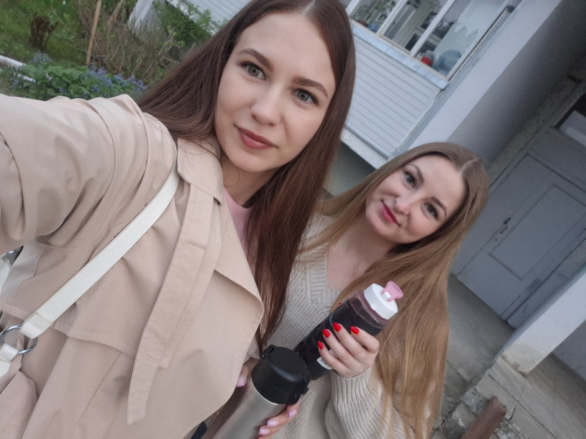 Two young women are posing for a selfie outdoors. The woman on the left has long, dark brown hair and is wearing a beige trench coat with a white strap bag. The woman on the right has long, blonde hair and is wearing a light-colored sweater. She is holding a black thermos and a water bottle filled with a dark-colored liquid. The background includes a building with light gray siding and windows, some greenery, and a partially visible door. The location is identified as Chaikovsky, Russia, based on additional available information.