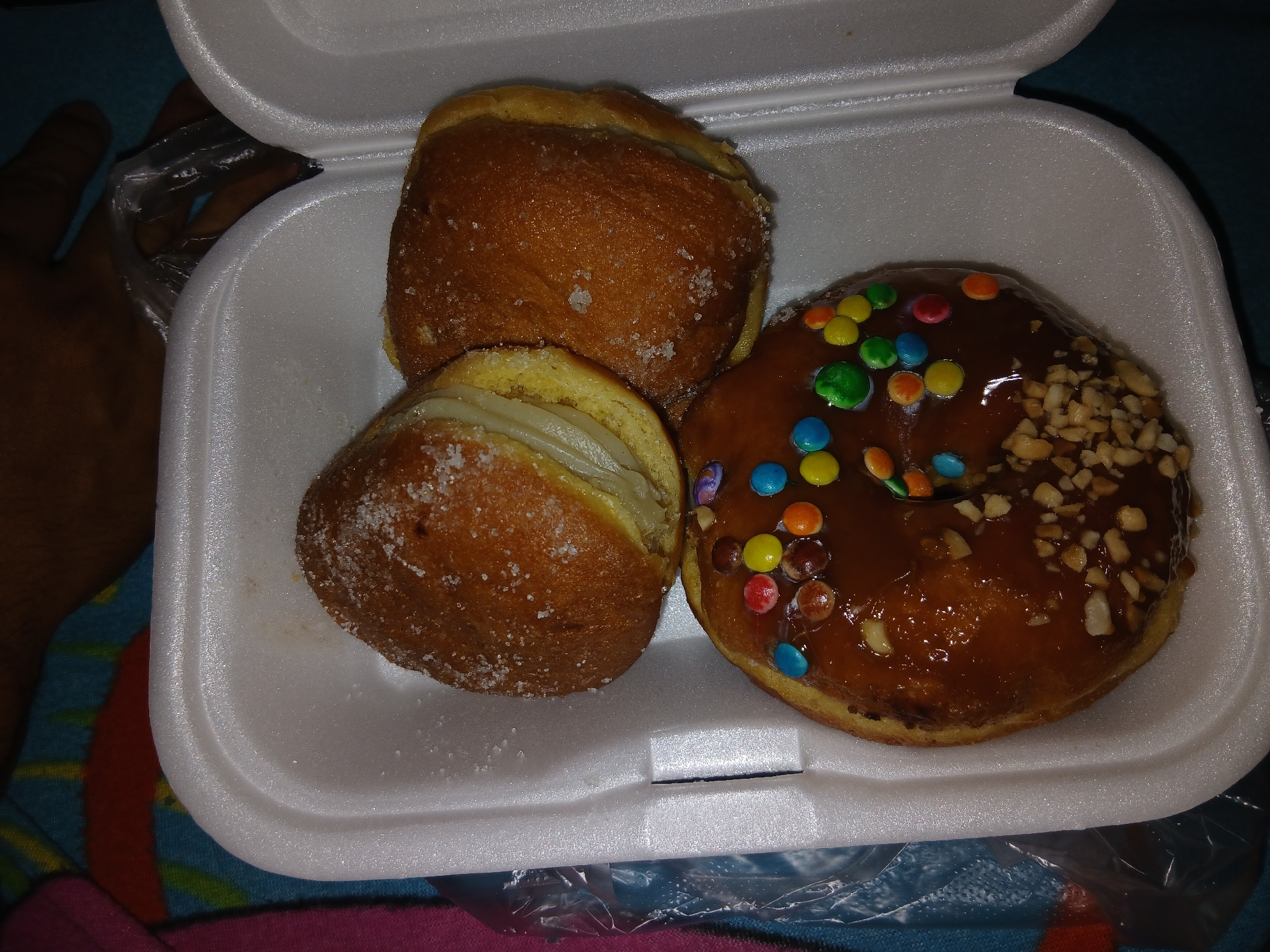 The image depicts three donuts in a white styrofoam container. Two donuts appear to be sugar-dusted and filled with cream. The third donut is glazed with chocolate, sprinkled with colored candies, and chopped nuts. The container is likely on a patterned fabric surface. A hand holding a plastic bag is partially visible in the upper left corner of the image. There is no other information that suggests this image was taken in the city of Altagracia, Venezuela.