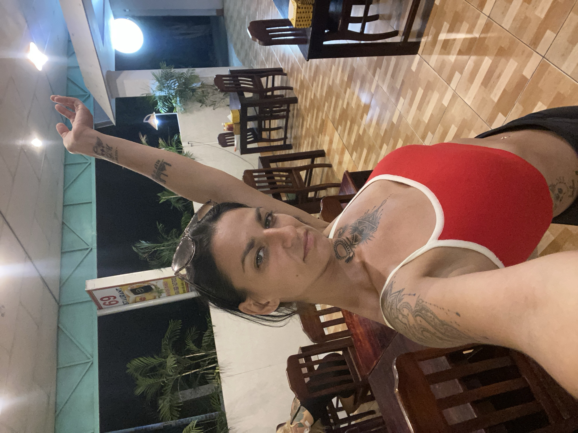 The media file shows a woman posing for a selfie in what appears to be a restaurant or dining area. The woman is in the foreground, looking at the camera with a neutral expression. She is wearing a red top with white trim and black shorts. She has several tattoos, including one on her chest and arm. Her sunglasses are perched on her head. The background shows tables and chairs, with a tiled floor. There are also some plants visible, and a sign that reads "69 RESTAURANT". The lighting suggests it is nighttime, possibly evening. The overall scene is casual, and the woman appears relaxed. The location cues like the "69 RESTAURANT" sign and the presence of tropical plants suggest a location in Phan Thiết, Vietnam.