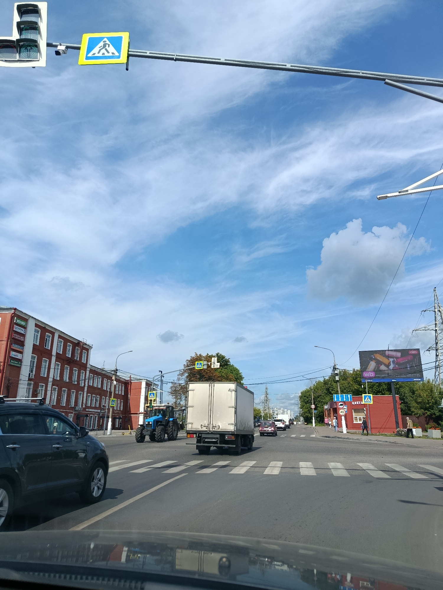 The image shows a street scene in Orekhovo-Zuyevo, Russia, on a day with a blue sky and scattered clouds. In the foreground, the dashboard of a car is visible, reflecting buildings and traffic lights. A dark-colored SUV is on the left side of the road, its front end partially visible.
Further down the street, a blue tractor is driving across a pedestrian crossing. To the right of the tractor is a white box truck. The road has clearly marked white pedestrian crossings.
On the left side of the image, a multi-story red brick building with many windows lines the street. Signs with Russian text are mounted on its facade.
On the right side of the road, a red building with a white window is visible, with people walking on the sidewalk in front of it. Above this building, a large billboard advertising "wb" (likely Wildberries) is displayed. Traffic lights and various road signs, including a pedestrian crossing sign, are present. Power lines and utility poles are also visible throughout the scene. Trees with green foliage line the street in the background.