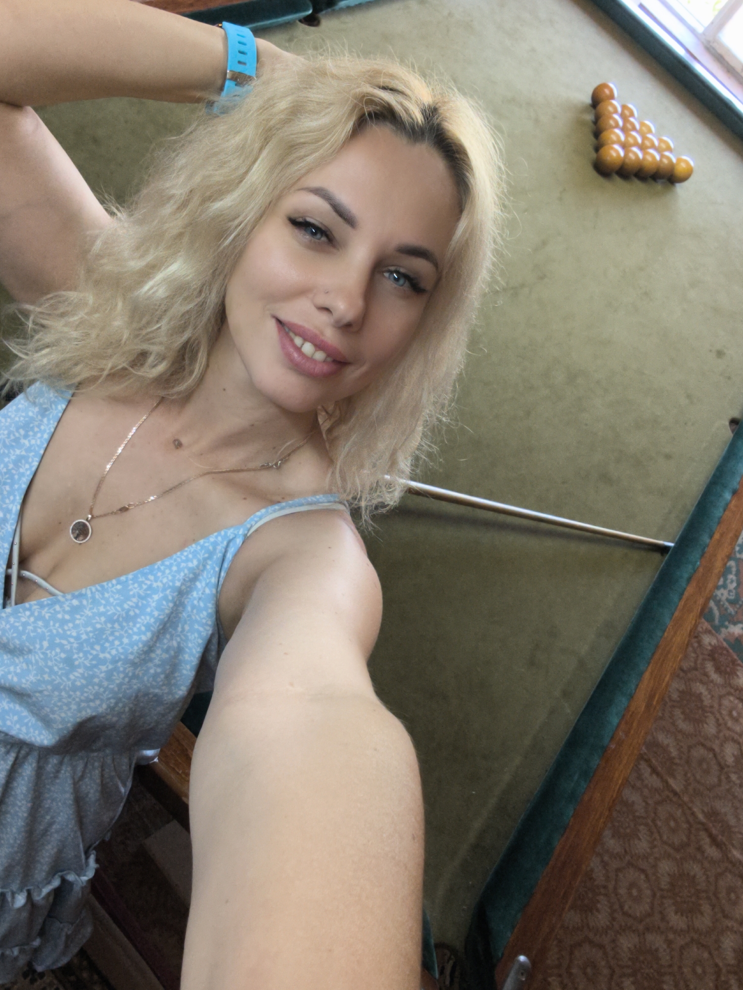 An adult female with long, wavy blonde hair and blue eyes smiles directly at the camera in a selfie captured in Терновский округ, Ukraine. She wears a light blue sundress with a white floral pattern, a V-neck, and thin shoulder straps. A thin gold-toned necklace with a circular pendant adorns her neck, and a bright blue fitness tracker is on her raised right wrist. Subtle winged eyeliner, mascara, and pink lipstick are visible, along with a small stud piercing on her left nostril and a small mole on her chest. Her right arm is bent and raised, with her hand likely behind or near her head, while her left arm is extended, holding the camera, with its forearm foreshortened in the lower center of the frame. Behind her, partially visible, is an indoor setting featuring a green-felted billiards table with dark wooden rails. On the far side of the table, 15 amber-colored billiard balls are racked in a triangle formation near a corner pocket. A long, slender silver-toned metal bar rests horizontally across the table's surface. Natural light enters from a window visible in the upper right. A brown and orange patterned rug is partially visible on the floor next to the table.