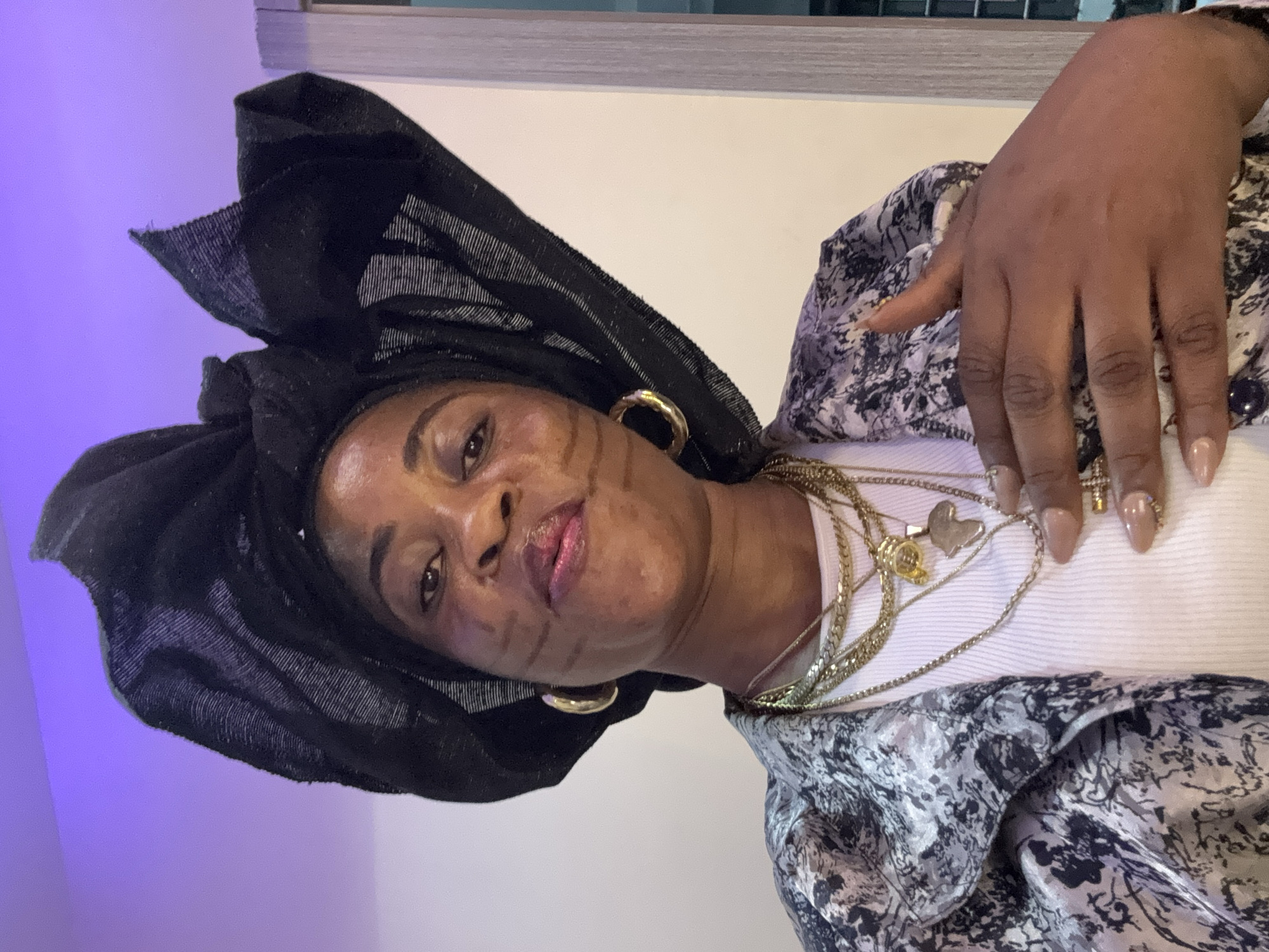 A medium shot captures a Black woman with dark skin wearing a large black headwrap and a patterned jacket over a white ribbed top. She has distinctive dark markings on her cheeks, large gold hoop earrings, and multiple gold necklaces, one with a pendant shaped like Africa and another with a cross. Her right hand is placed over her chest, revealing manicured nails. The background consists of a wall with subtle lighting creating a purple hue at the top and a lighter tone below, with a hint of a vertical structure on the right. The scene is indoors, likely a room, with no clear indication of the city of Ajara, Nigeria.