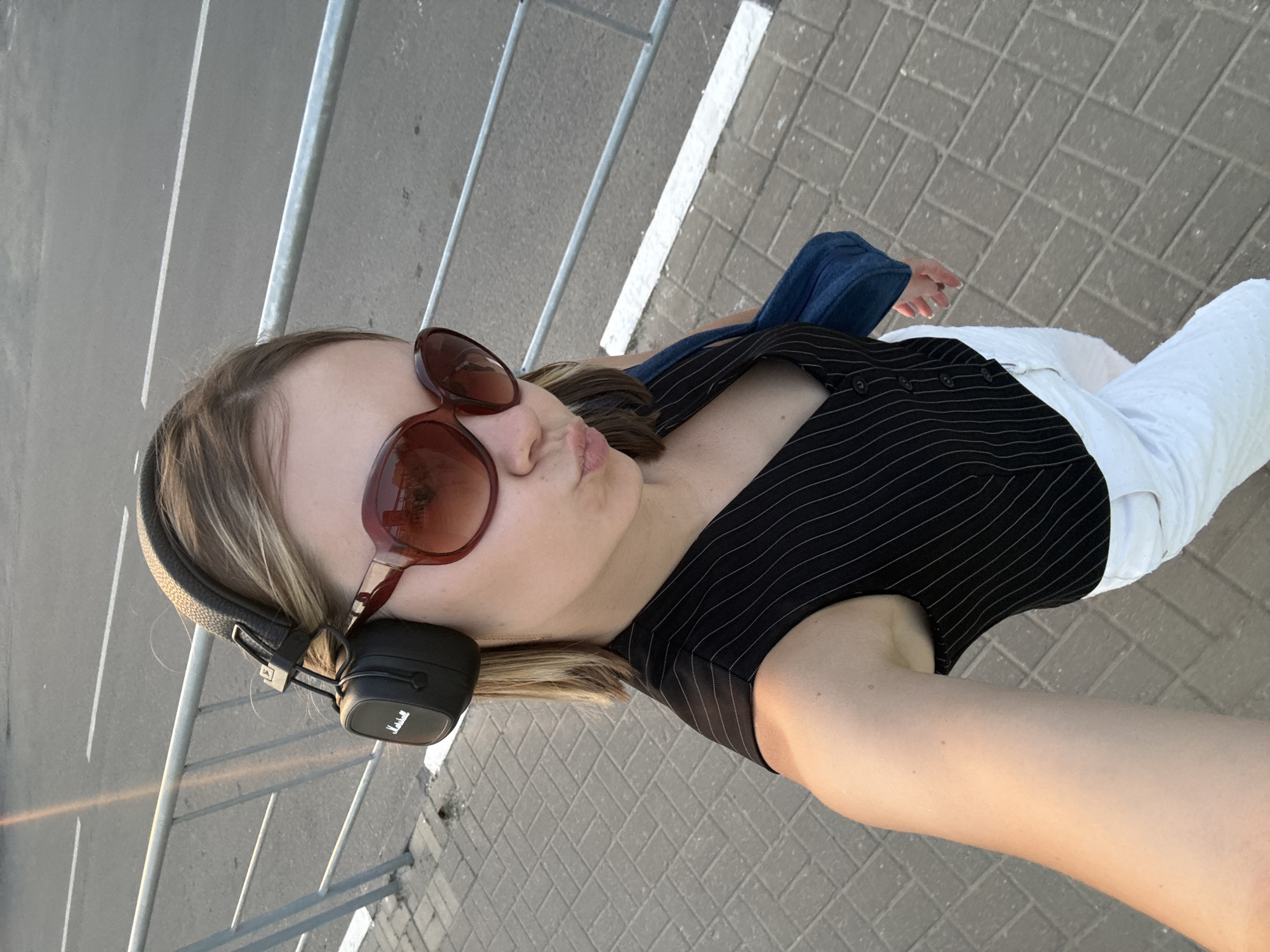 A young woman is standing outside on a brick-paved surface near a metal railing in Severnyi, Russia. She is wearing a black and white striped top, white pants, and has a blue jean jacket draped over her shoulder. Sunglasses are perched on her face and black Marshall headphones are around her neck. She is pouting her lips and posing for the selfie.