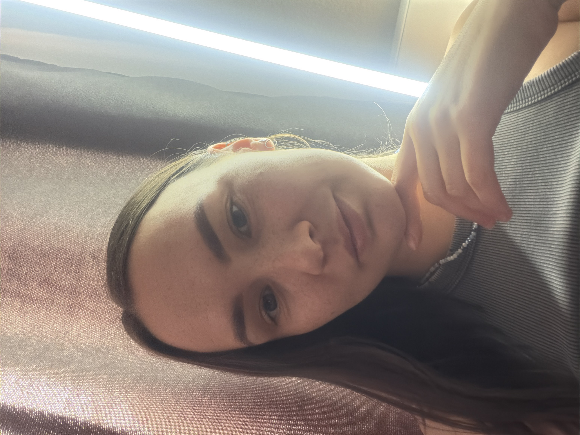 A young woman with brown hair and fair skin is positioned in the frame, her face close to the camera. Her dark hair falls to her right, partially obscuring her shoulder and a gray striped shirt. She has freckles and her hand is near her neck. Above her, a bright linear light source illuminates the scene. The background consists of a textured surface, possibly fabric, with a metallic sheen. The photograph's orientation is slightly tilted. There is no information to determine the location to be Chaikovsky, Russia.