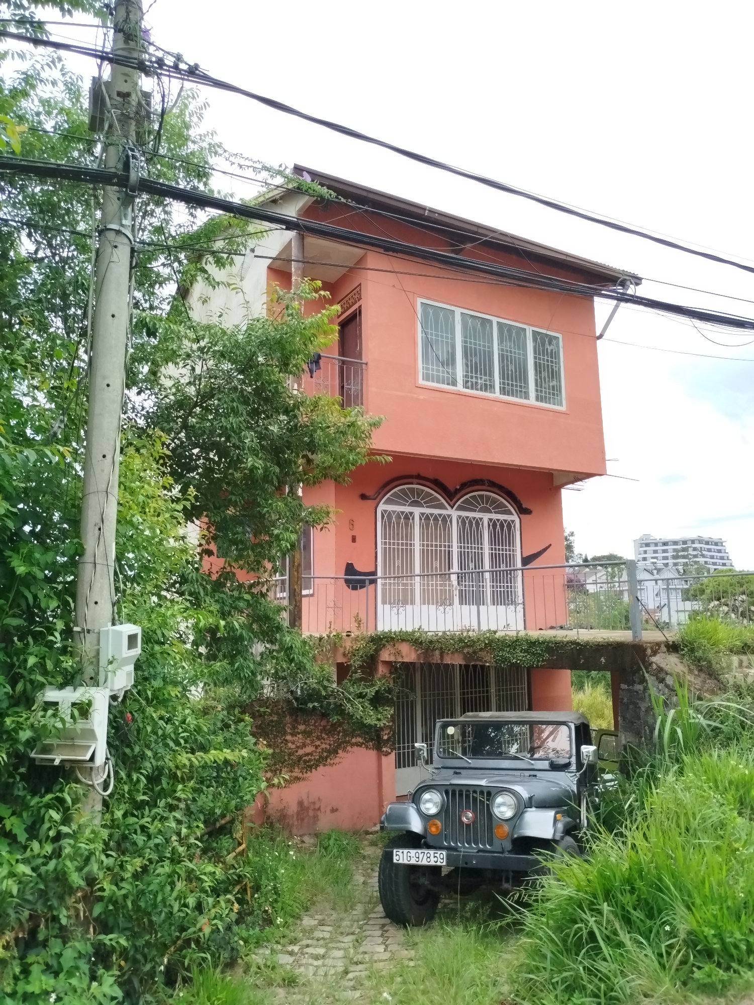 A three-story, peach-colored building with white window frames and wrought-iron balconies sits on a hillside. A gray jeep with license plate "51G-978 59" is parked in front of the building. Overgrown foliage surrounds the building and the jeep. Utility poles with multiple wires extend into the frame on the left and across the top of the image. The location is consistent with an area in Da Lat, Vietnam. No people are visible in the scene.
