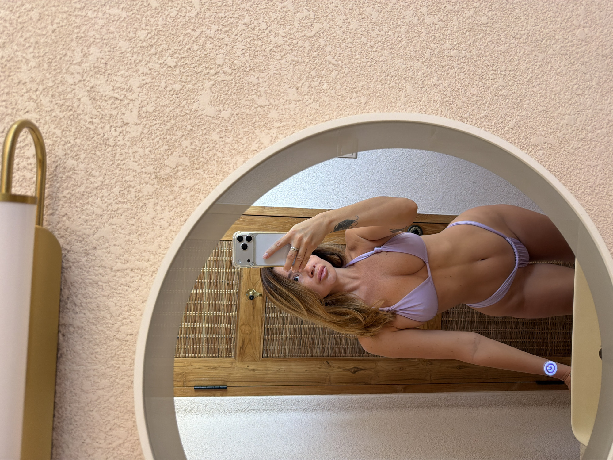 A woman is depicted taking a mirror selfie, reflected in a large, circular mirror with a light-colored frame. She is holding a modern smartphone in a clear case, partially obscuring her face, though her full lips and long, light brown hair are visible. She is wearing a light purple two-piece bikini, showcasing her toned physique. A small tattoo, appearing to be a feather or wing, is visible on her left forearm. On her right wrist, she wears a circular device with a glowing blue-purple symbol, possibly a power icon.
The mirror is mounted on a textured, light beige wall. To the left of the mirror, a golden, curved light fixture with a white shade is partially visible. The reflection in the mirror reveals a room with a prominent wooden background, featuring a woven pattern that resembles rattan or bamboo matting, suggesting a door or cabinet. A dark handle or knob is visible on this wooden surface. Below the wooden element, a light-colored, textured surface, likely the floor or a lower wall, is reflected. The setting appears to be an indoor space in Mengwi, Indonesia, possibly a hotel room or resort bathroom, with natural lighting.