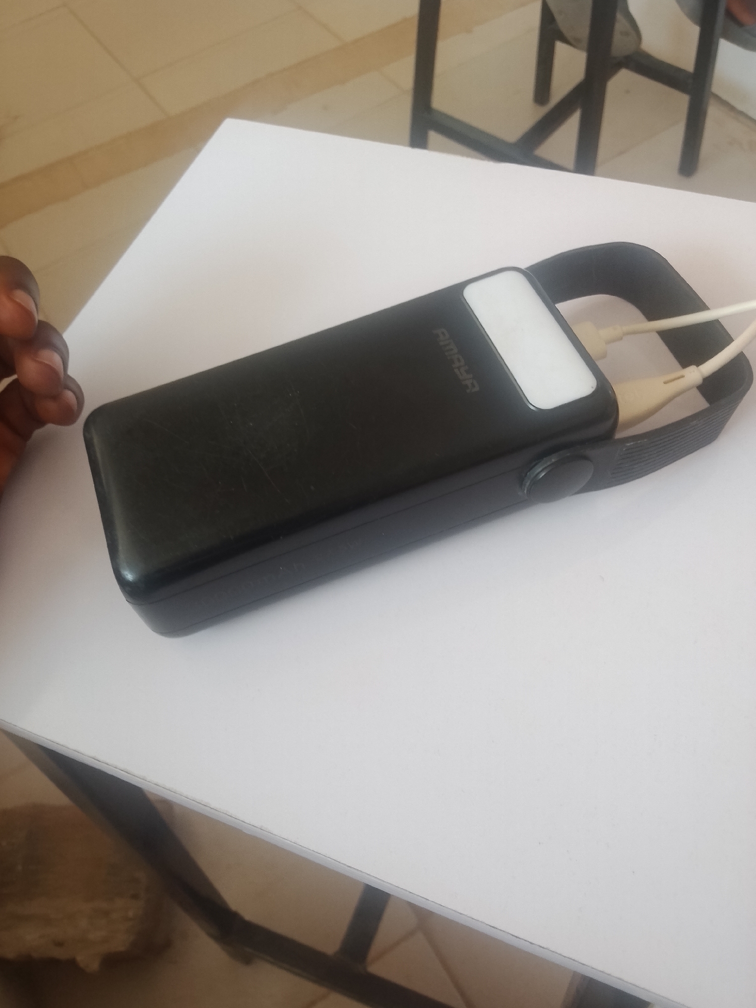 A black power bank with a white band and a grey strap is resting on a white surface. Two white charging cables are plugged into the power bank. The power bank has the brand name "AMAYA" printed on it. The scene is indoors, possibly in a classroom or office, with a tiled floor and furniture visible in the background.
