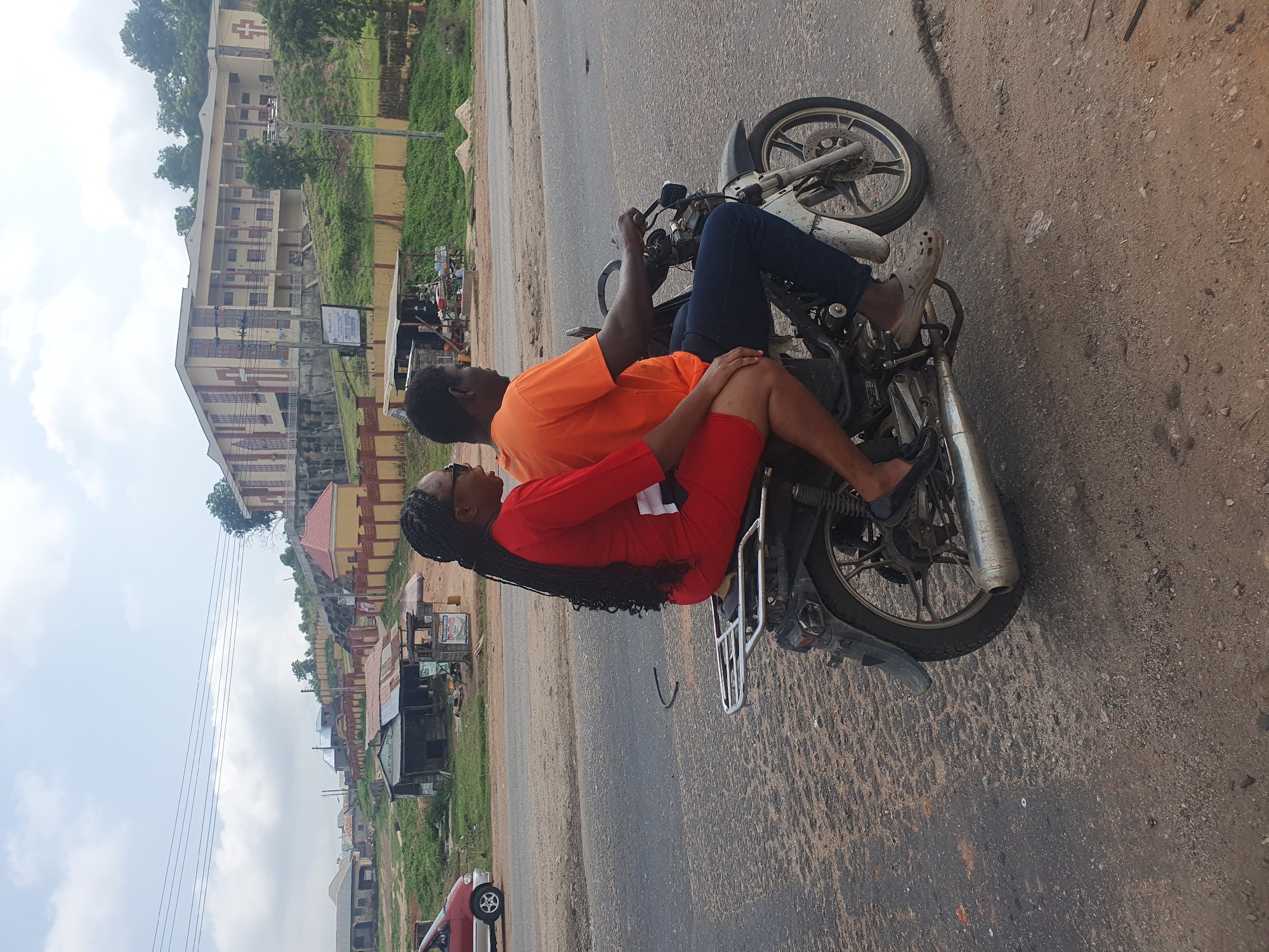 An overcast day in Lokoja, Nigeria, finds two individuals on a motorcycle traveling on a paved road. The person in the front, a woman with dark, braided hair, wears a vibrant red dress. The individual behind her, a man with dark skin, is dressed in an orange t-shirt and dark blue pants. They are moving from left to right across the frame, with the motorcycle's rear wheel in the foreground. The environment includes a roadside with scattered debris and vegetation. In the background, various structures are visible, including a large, multi-story building on a hill, residential housing, and small commercial stalls. A red car is parked on the left side of the road.