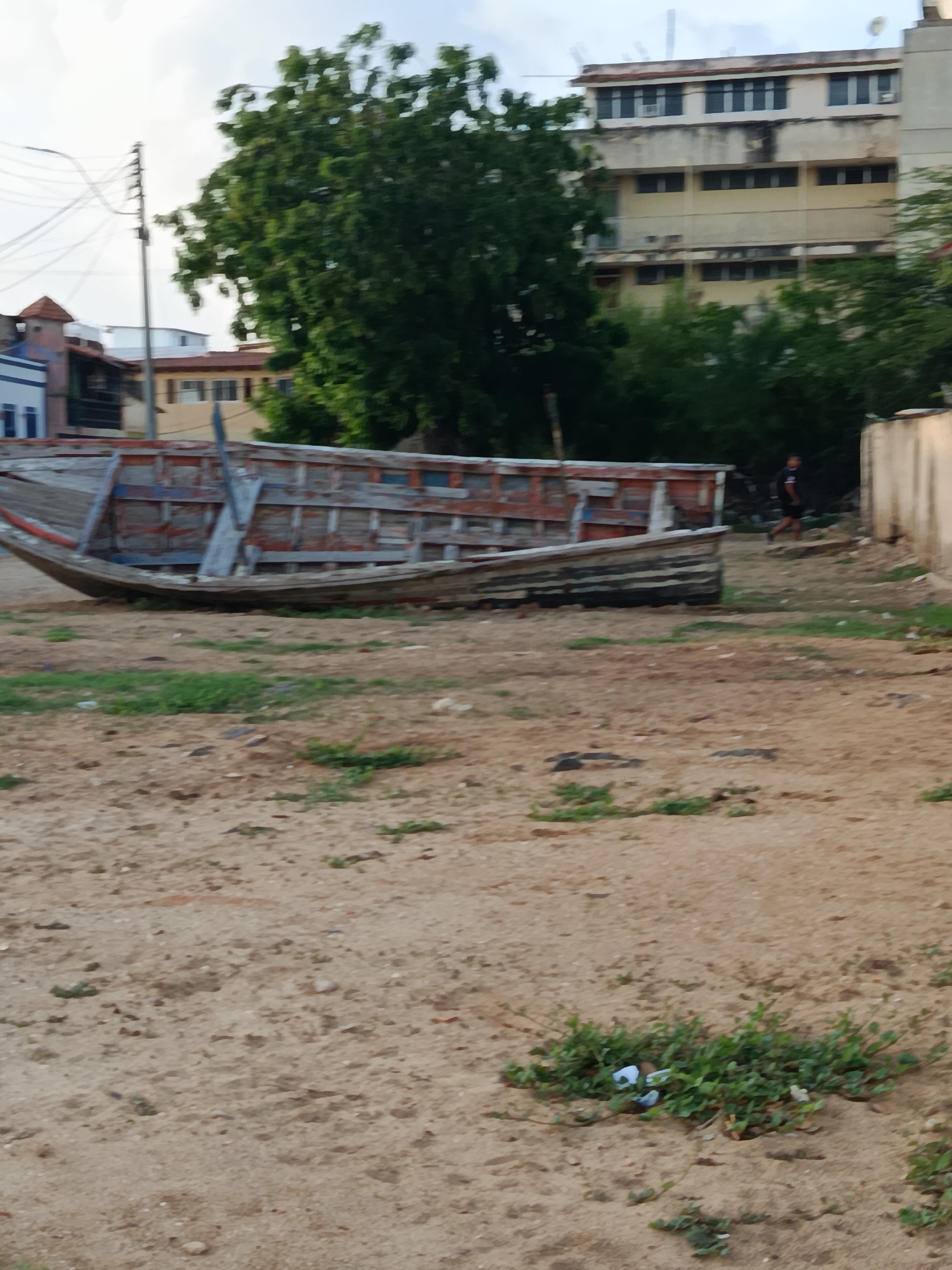 An abandoned wooden boat is situated on a sandy, uneven terrain with sparse vegetation in the foreground. The boat, weathered and aged, displays remnants of red and blue paint. In the background, a multi-story building with numerous windows stands behind a dense cluster of trees. To the right of the boat, a light-colored wall is visible, and a person in dark clothing is walking away from the boat. The sky is overcast. The location is identified as Juan Griego, Venezuela.