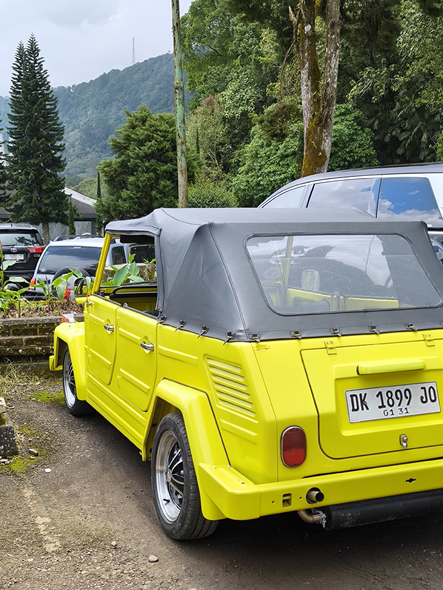 A yellow Volkswagen Thing, license plate DK 1899 JO, is parked in an outdoor area in Wanagiri, Indonesia. The car has a black convertible top, chrome rims, and is oriented towards the right. It's parked on a gravel and dirt surface. Other cars are parked in the background, along with some greenery including various trees. A mountain range is visible in the distance under a cloudy sky.