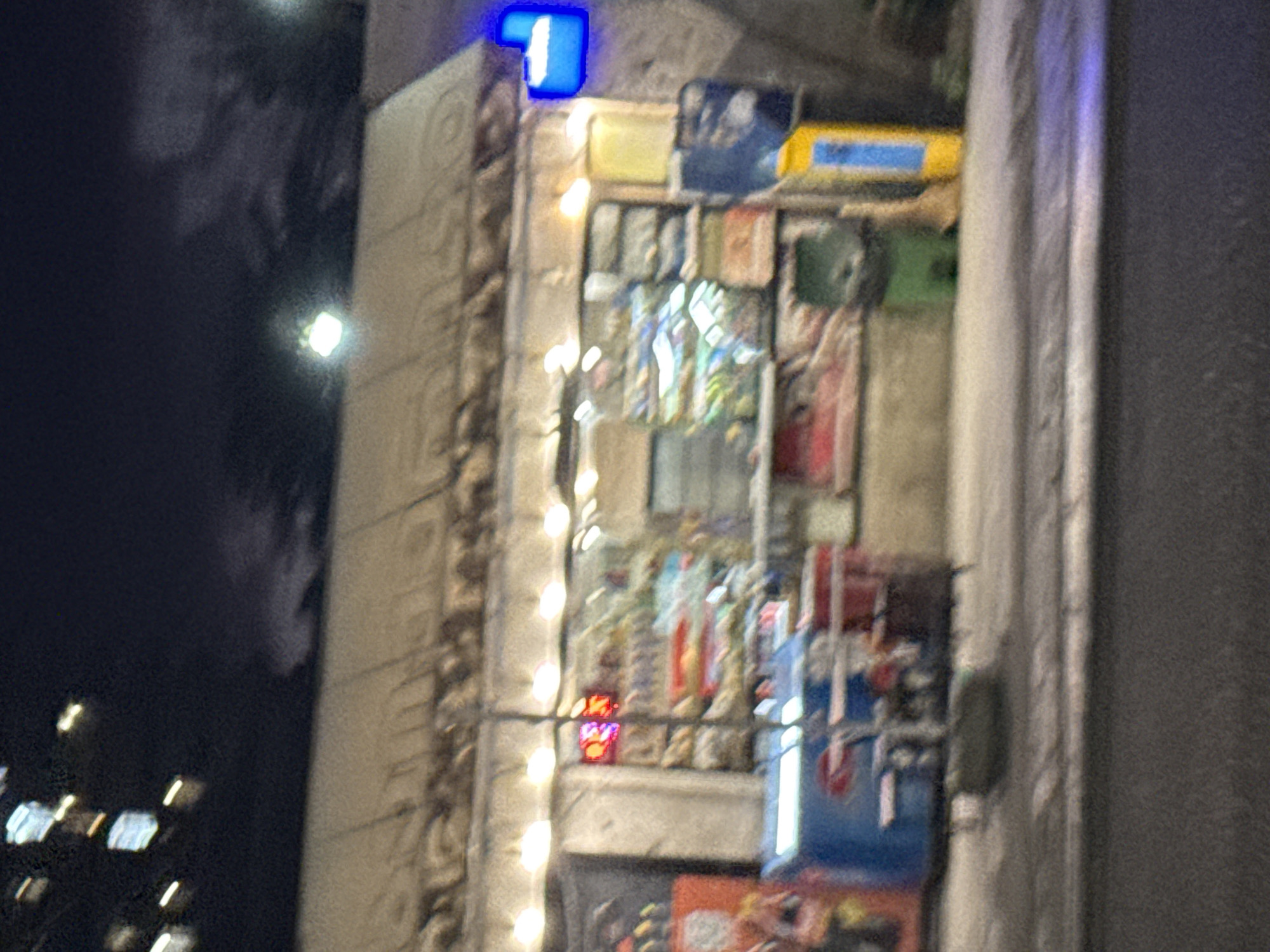 The image shows a night view of a small storefront, possibly a convenience store or kiosk, in Tbilisi, Georgia. The facade of the building is covered by a canvas awning illuminated by string lights. Inside, shelves are stocked with various items, some of which appear to be food or drinks. In front of the store, a blue refrigerated display case with a Pepsi logo is visible. To the right of the storefront, a blue and yellow sign or trash receptacle stands on the sidewalk. The street in front of the store is paved and appears to be dimly lit. The overall impression is of a typical urban commercial establishment operating at night.