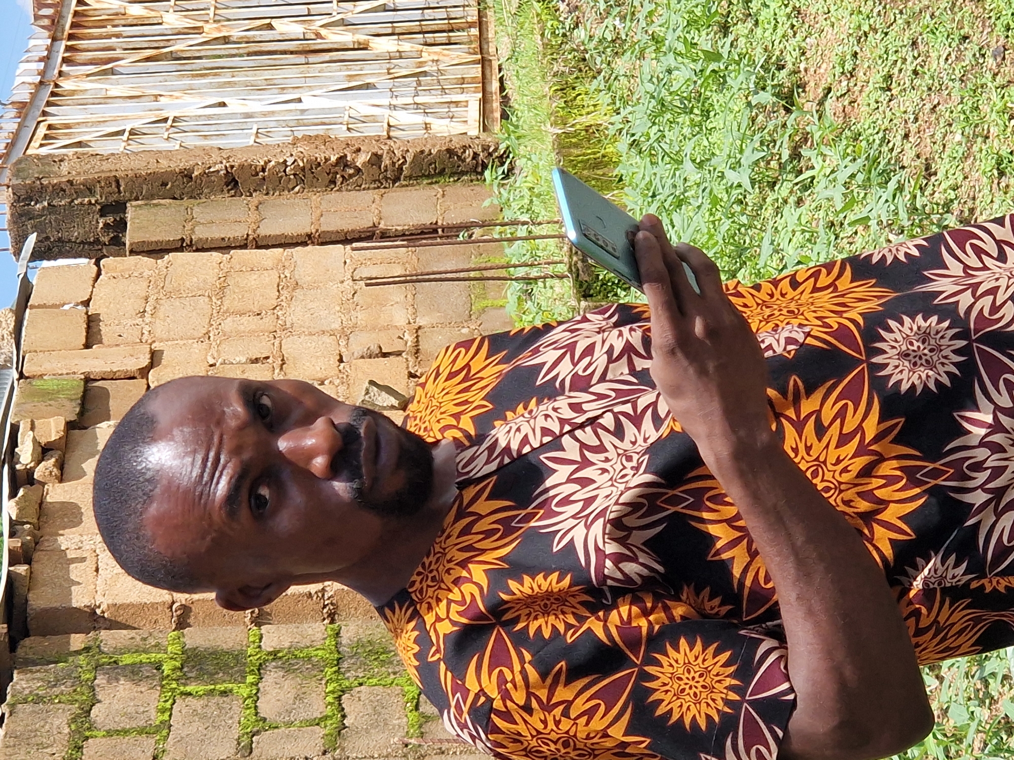 A Black man stands in front of a brick wall and a metal gate. He is wearing a black shirt with a yellow and red floral pattern. He is holding a light blue smartphone in his right hand. He has short dark hair and a goatee. The wall is made of light brown bricks. The gate is made of white metal bars. There is green foliage in the background. The image is taken outdoors in daylight.