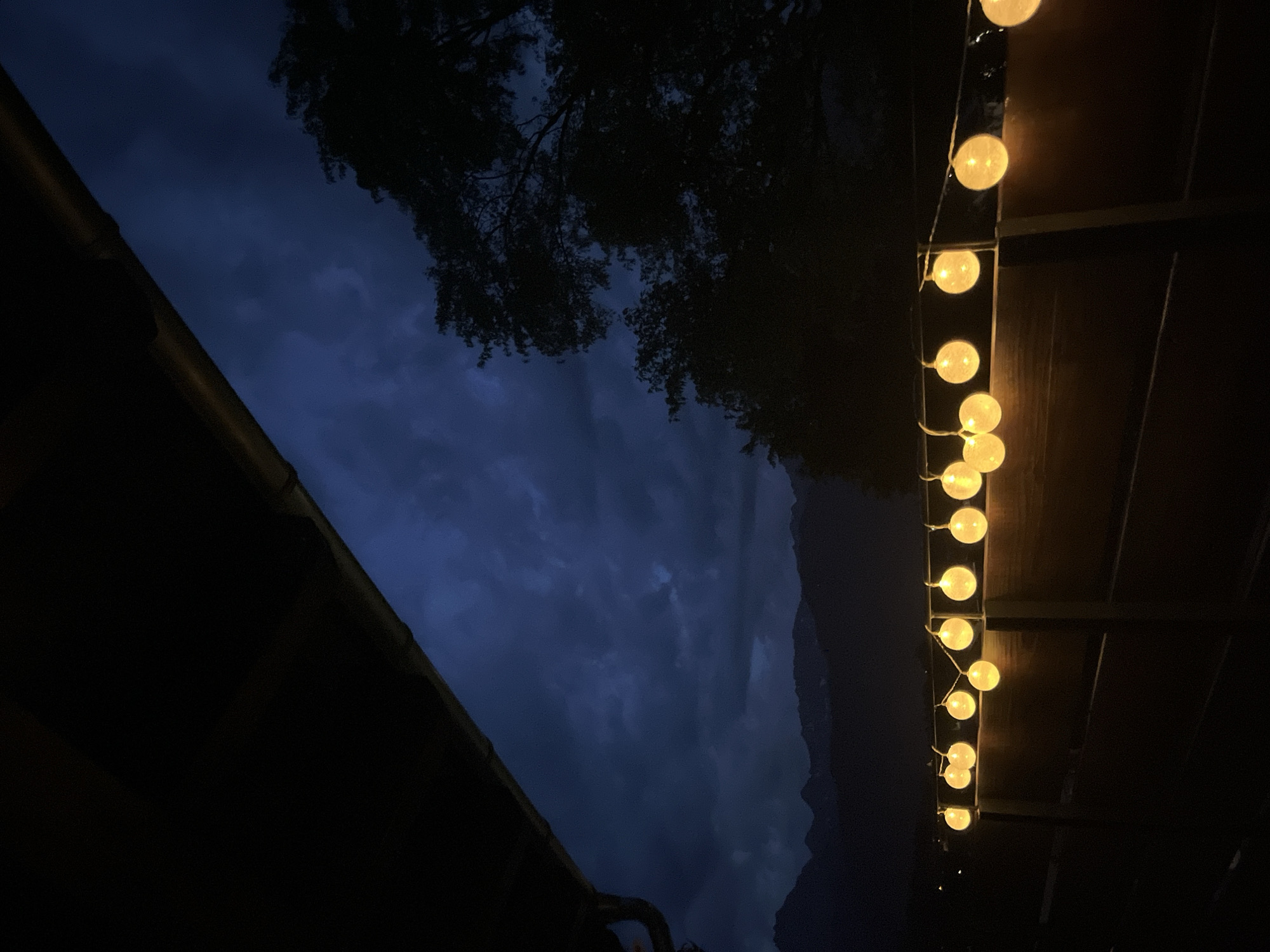 The image depicts a dimly lit outdoor scene, likely at dusk or night, with a dark, cloudy sky overhead. In the foreground, a string of illuminated round lights is draped along the top of a wooden fence. The lights are a warm yellow color and are spaced evenly. The fence itself is dark and appears to be made of horizontal wooden planks. Above the fence, silhouetted trees are visible against the dark sky. In the far background, a mountain range is faintly discernible. The perspective suggests the viewer is on a balcony or raised platform, as indicated by the dark overhang in the upper left corner of the image. The location is specified as Garmisch-Partenkirchen, Germany, suggesting a mountainous or alpine setting. There are no people or animals visible in the image.