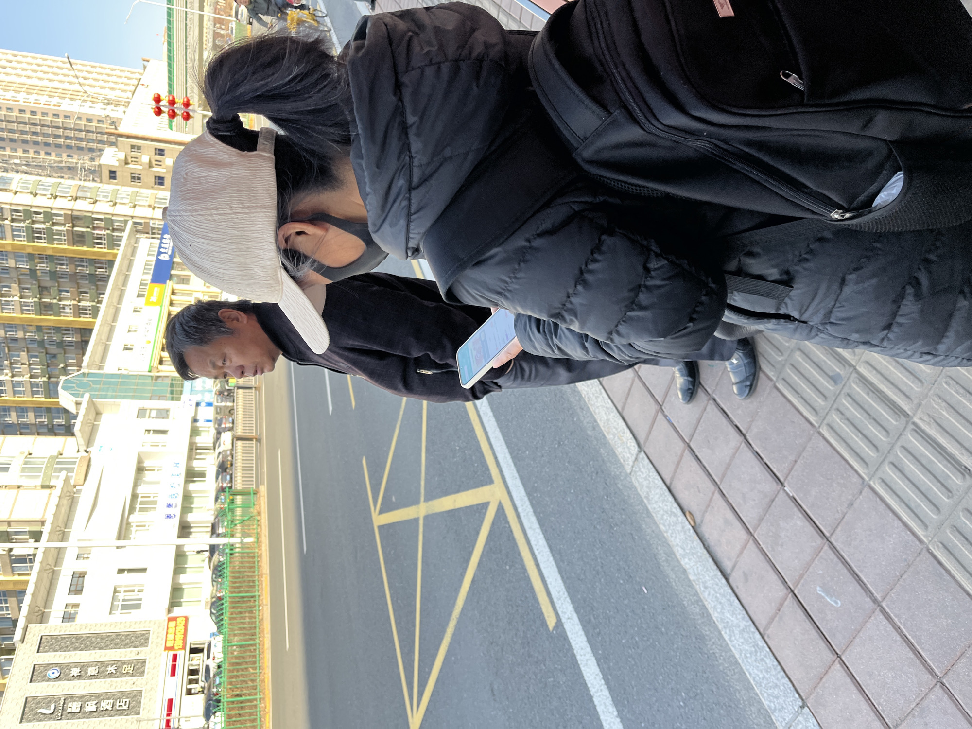 A medium-angle outdoor shot shows two people standing on a sidewalk in what appears to be Changchun City, China. The person closest to the camera is wearing a black puffer jacket and a black backpack. They are also wearing a white baseball cap and a black face mask. In their hands, they hold a smartphone. The other person, slightly behind and to the left, is wearing a black jacket and holding a smartphone. The background shows a street with yellow lines on the asphalt, a sidewalk with tactile paving, and buildings. A sign in Chinese characters is visible on one of the buildings. Traffic lights and other signs are also present in the distance. The shot is slightly tilted counter-clockwise.