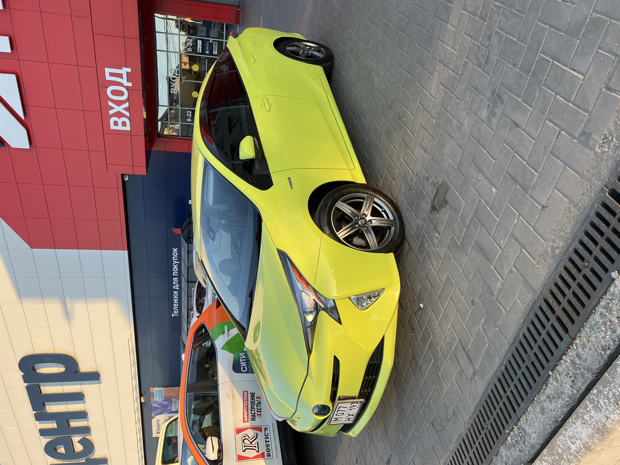 This media file is a rotated, eye-level, full-shot photograph of a bright yellow Toyota Prius parked on a sidewalk outside a retail store. The store has a red and dark blue facade. Large white lettering on the red section spells out "ВХОД" (Entrance), and smaller text on the dark blue section also says "ВХОД" with operating hours "8-22" displayed twice. Next to the "ВХОД" sign, text on a dark band reads "Тележки для покупок" (Shopping carts).
The car's front passenger side is prominently featured, showing its distinctive headlights and grille. The car's license plate is visible and reads "M077 HX 193". To the left of the yellow Prius, other vehicles are partially visible, including a white car with orange and green graphics and the text "СИТИ" (City) and "МАРШРУТ ПОСТРОЕН НАСТРОЕНИЕ ЕСТЬ!" (Route built, mood is there!). Another car has a logo that appears to be "R ROSTIC'S". Large grey letters spelling "центр" (center) are on the building to the left.
The sidewalk is paved with dark gray interlocking bricks laid in a herringbone pattern. A black metal grate, likely a drain cover, runs along the right edge of the frame. The scene appears to be outdoors during the daytime, with sunlight casting shadows, suggesting it is late afternoon or early evening. The lighting on the car indicates it is a sunny day. The overall setting is a commercial area, likely in Krasnodar, Russia, given the Cyrillic text. No people are visible in the photograph.