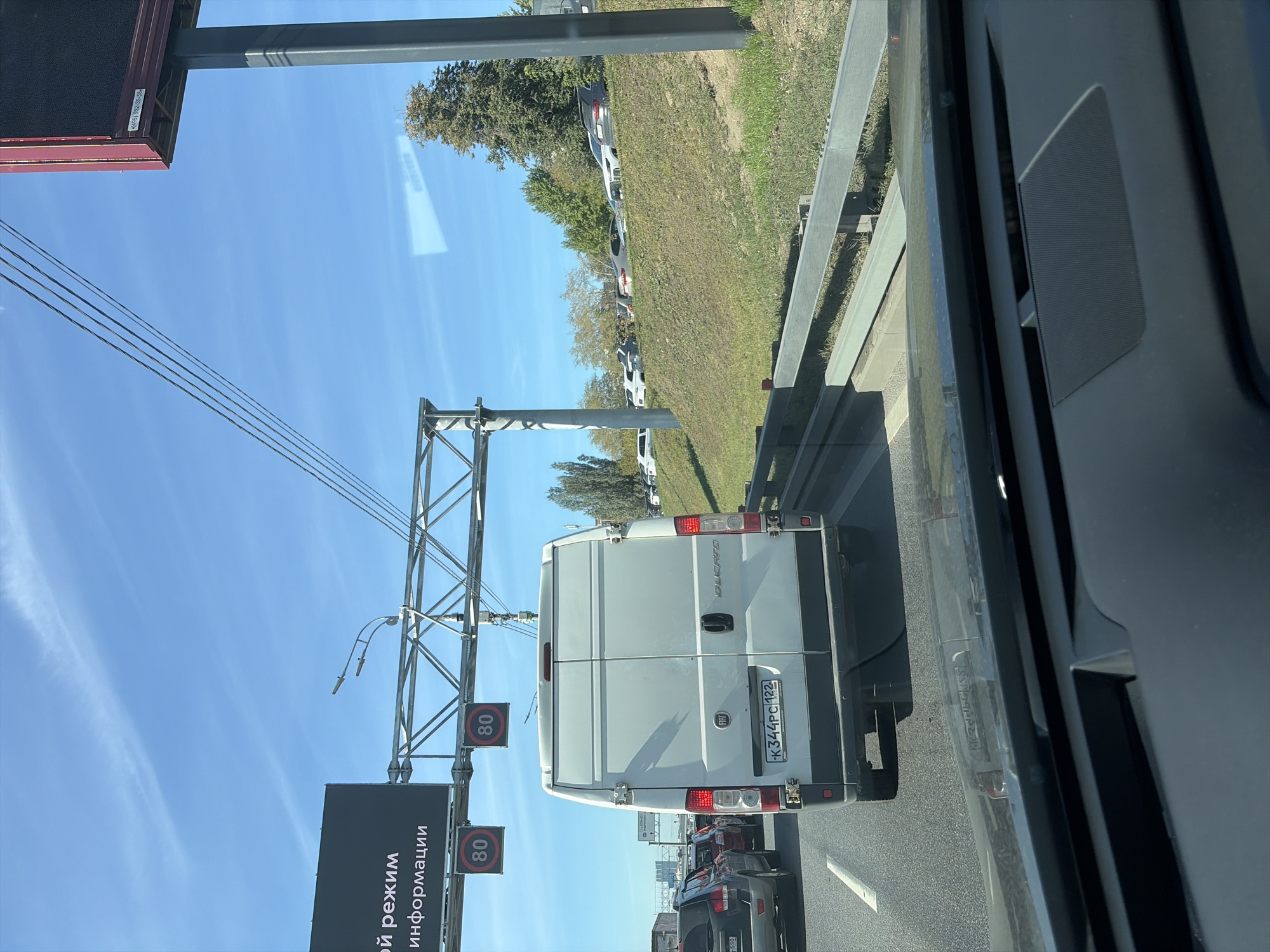The image is a street-level view of a highway with multiple lanes of traffic. A white Fiat Ducato van is in the foreground, traveling in the right lane. The van has a license plate that reads "K344PC 122". Two speed limit signs displaying "80" are visible overhead, indicating a speed limit of 80 kilometers per hour. On the left side of the highway, there is a dark billboard with Cyrillic text. On the right side of the highway, there is a grassy embankment with several cars parked on it, and beyond that, trees. The sky is blue with some wispy clouds. The overall scene suggests a sunny day with moderate traffic. Based on the license plate format (K followed by three digits and two digits with the region code 122, which corresponds to Kostroma Oblast, though similar plates can be seen in other regions) and the general appearance of the road and surrounding infrastructure, this scene is likely in or around Moscow, Russia, a major urban center known for its extensive highway network and diverse vehicle population.