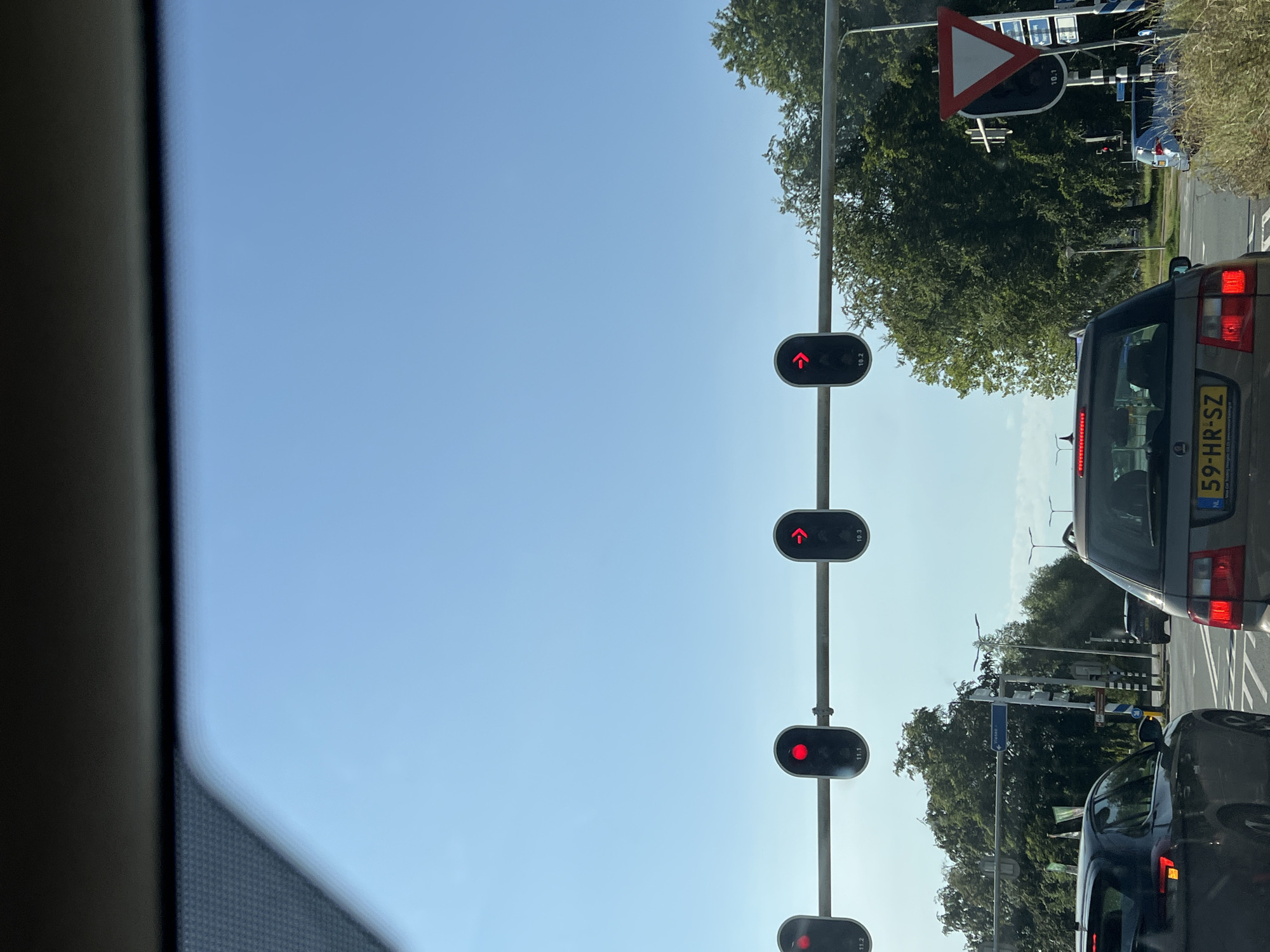 The image shows a view from inside a car, looking forward at a traffic light intersection on a clear, sunny day. The sky is a vibrant blue with no clouds visible. In the foreground, the dashboard and windshield of the car are visible at the top.
The main focus of the image is a series of traffic lights suspended over the road. There are four traffic lights visible in a horizontal row. Each light is red, indicating that traffic should stop. The first light from the left shows a standard red circle. The second light from the left also shows a standard red circle. The third light from the left displays a red arrow pointing to the right. The fourth traffic light, furthest to the right, also shows a red arrow pointing to the right. Some numerical labels are visible beneath each traffic light.
Below the traffic lights, several cars are stopped, waiting at the intersection. The car directly in front of the viewer is a dark-colored hatchback. To the right of this car is a silver station wagon with a license plate that reads "59-HR-SZ". Further down the road, other vehicles and traffic signals are visible.
On the right side of the road, a yield sign is prominently displayed, a red triangle with a white center and a black border. Beyond the intersection, lush green trees line the sides of the road, indicating a somewhat suburban or park-like setting. The overall scene depicts a common urban or suburban traffic situation, with cars stopped at a red light.