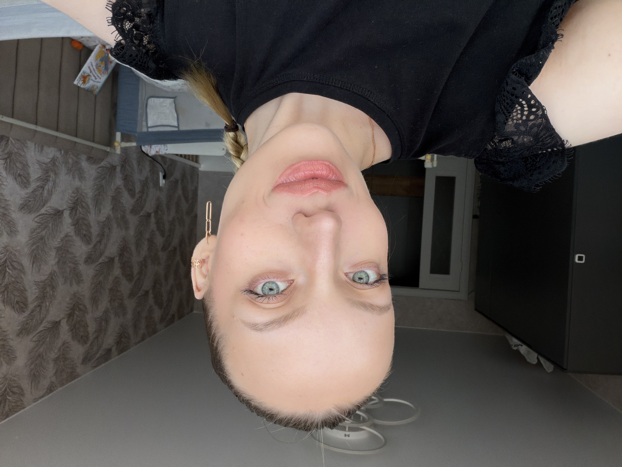 The image displays a headshot of a person, likely female, taken indoors, with the image itself oriented upside down.
The subject has fair skin, light-colored eyes (appearing blue or green), and light-colored hair (blonde or light brown) which is styled with a braid visible on the right side of the frame (when the image is viewed upright). Her eyebrows are well-defined, and her eyelashes are emphasized with mascara. Her lips are coated with a pinkish-nude, glossy lipstick. She wears a black top, possibly a t-shirt, which features a detailed black lace trim on the short sleeves or shoulder area. A thin, gold-toned chain necklace is visible around her neck. On her left ear, a delicate gold-toned ear cuff or earring, possibly in a leaf or feather design, is worn. The person is looking directly into the camera with a neutral expression.
The background behind the person indicates an indoor room. To the left side of the frame (which would be the right side if the image were upright), there is a wall covered in wallpaper featuring a repeating pattern of dark, stylized leaf or feather motifs on a grey background. A power outlet is visible on this wall. The floor appears to be a uniform dark grey.
In the upper left corner of the frame (bottom right when upright), there is furniture, possibly a bed or sofa, covered with a light-colored fabric. An open book with a colorful illustration on its cover is resting on this furniture, and a small, round orange object (possibly a fruit or toy) is also visible. Further in the background, a dark grey or black piece of furniture, resembling a cabinet or wardrobe, is partially visible, along with what appears to be a white door frame or window. A circular, white light fixture, possibly a ring light or ceiling light, is partially visible near the bottom edge of the image (above the person's head when upright).
No clearly discernible text is visible in the image, although the book cover has illustrations and faint text that cannot be read.