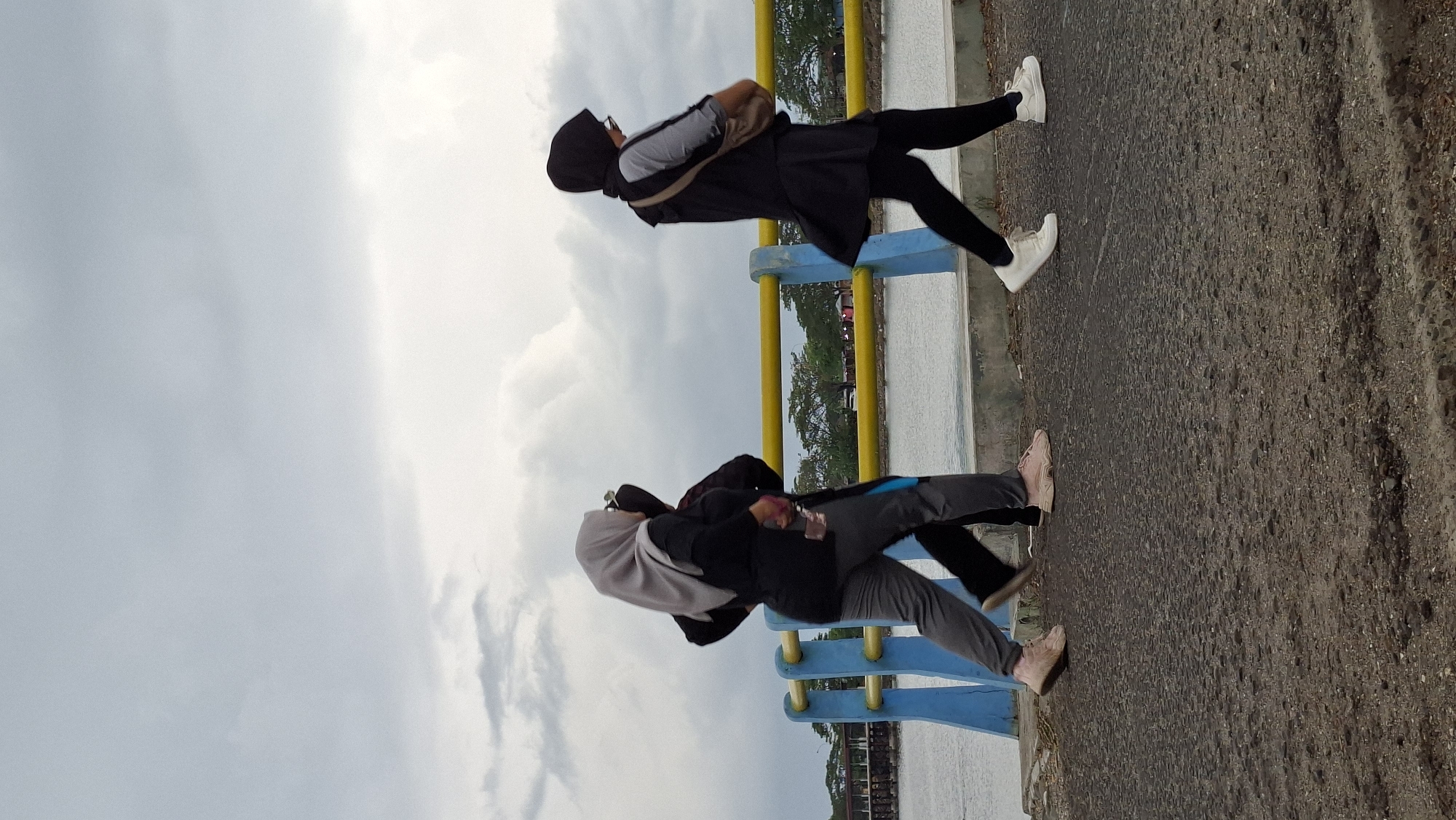 The image shows three individuals walking on a paved walkway next to a body of water. A railing with yellow and blue posts separates the walkway from the water. In the background, trees and some buildings are visible. The sky is cloudy and overcast.
Specifically, this scene appears to be the Krueng Geukueh historical site in Lhokseumawe, Indonesia. The walkway is likely part of the walking path along the coast, offering views of the sea and the surrounding natural landscape. The railing is a common feature for safety and aesthetics in such public areas. The presence of individuals engaged in walking suggests a leisure activity, possibly for exercise or recreation. The overcast sky indicates a typical weather condition for the region, which experiences a tropical climate.