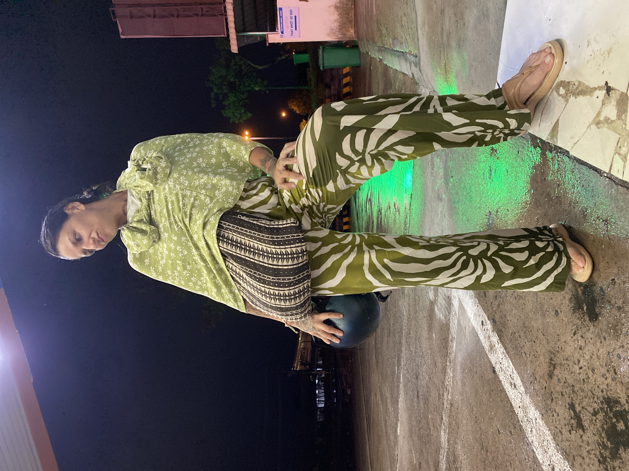 This nighttime image features a person standing outdoors in Phu Thuy Ward, Vietnam. The individual is positioned sideways, looking towards the camera with a neutral to slightly pensive expression. They are dressed in a light green long-sleeved top adorned with small white floral patterns, layered over a black and white patterned skirt or wrap. Their olive green wide-leg pants display a distinct white abstract or leaf-like design, and they are wearing light tan platform sandals. One hand, which is tattooed, holds a dark, possibly black, motorcycle helmet, while the other rests near their hip.
The scene is an urban or semi-urban setting, likely a service station or a well-lit commercial area. The dark sky confirms it is night. The ground, appearing to be concrete or pavement, is wet and reflective, with areas illuminated by a vibrant green light. In the background, a green trash can and part of a building are visible. A white sign is prominently displayed, bearing the "PETROLIMEX" logo and Vietnamese text that reads "GIỜ MỞ CỬA 24/24" (Open 24/24) and "ĐÊM THAY NHỚT XE MÁY" (Night motorcycle oil change). The wet surfaces and artificial lighting contribute to a nocturnal atmosphere.