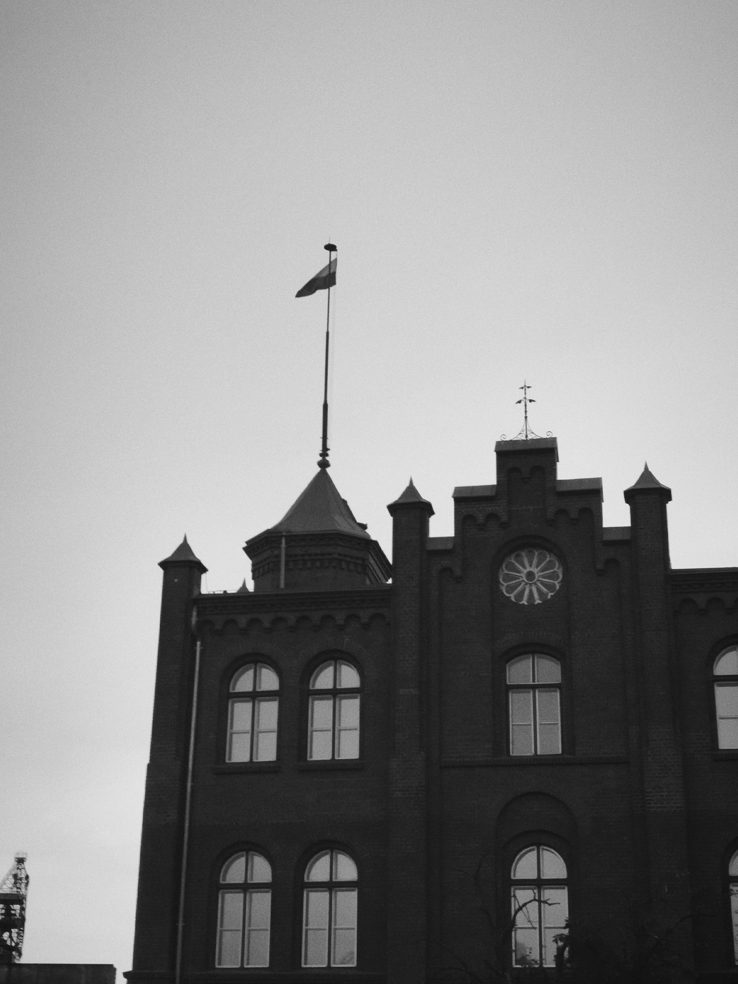The photograph, captured in black and white, depicts the upper section of a brick building, likely a historical structure, under an overcast sky. The building features arched windows with mullions and a decorative circular window with a geometric pattern in the center of its facade. A flagpole with a triangular flag is visible atop a turret on the left side of the building, and a cross-like ornament adorns the central peak of the roof. In the far left background, a portion of a metal structure, possibly a crane or industrial element, is discernible. The overall ambiance is somber and architectural, emphasizing the building's facade and its upper features against a muted sky. This building is the Old Brewery, located in the Wrzeszcz district of Gdańsk, Poland.