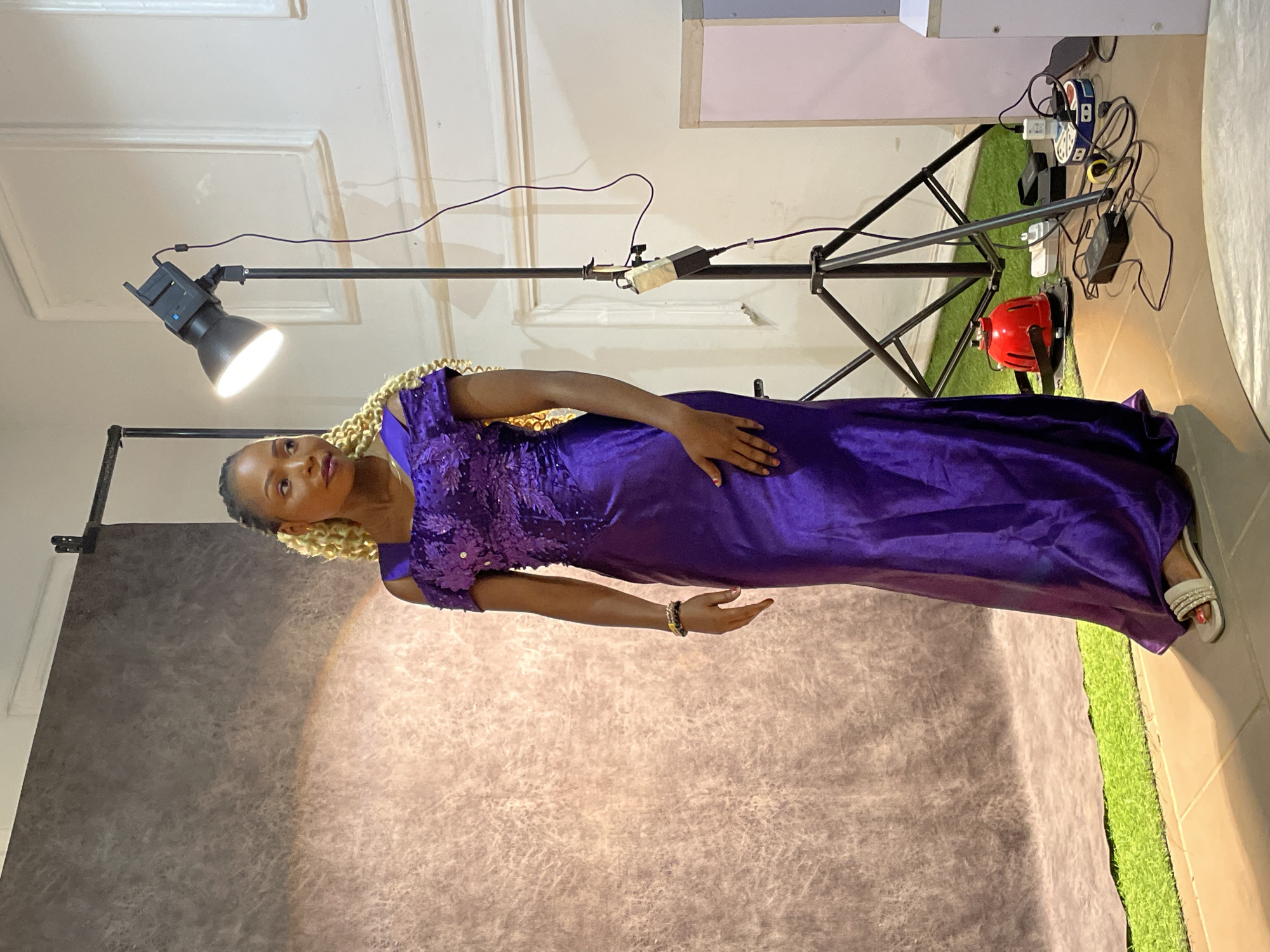 An indoor portrait features a woman posing in what appears to be a makeshift or small photo studio, likely in Barakin Akawo, Nigeria. The woman, with a medium skin tone and blonde braided hair styled in locs or twists, stands wearing a long, floor-length, deep purple dress. The bodice of her dress is intricately embellished with sequins or beads, while the skirt is made of a smooth, flowing fabric. She accessorizes with a dark-colored bracelet on her left wrist and wears white, open-toed sandals adorned with rhinestones. Her gaze is directed slightly upwards and to her left, with a composed expression.
The studio setup includes a textured grey and brown backdrop on the left side, providing a neutral background for the portrait. To the right, a strip of artificial green grass adds an unexpected pop of color and texture to the floor. Lighting equipment is visible, including a black light stand with a bright spotlight illuminating the woman from the left. Another light stand extends across the upper right portion of the frame. Various cables, a white power strip, and a red industrial-style fan are clustered on the artificial grass mat, indicating a functional, rather than pristine, studio environment. The walls in the background are white with some simple architectural molding, and there's a light-colored panel attached to the upper right wall. No visible text is present in the image.