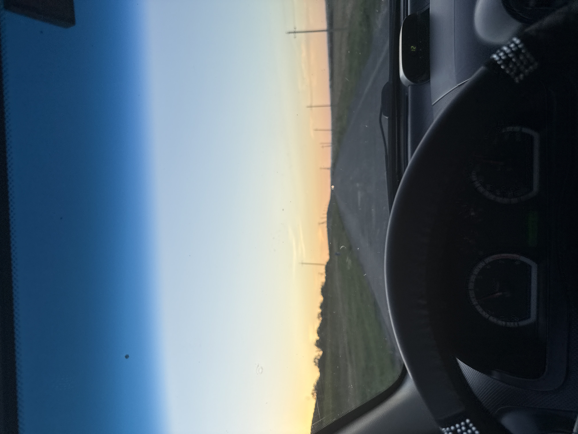 The image shows a view from inside a car looking out through the windshield. The car is driving on a rural road at dusk. The sky is a gradient of deep blue at the top to orange and yellow near the horizon, indicating sunset. The road is paved and stretches into the distance, with telephone poles lining the sides. The landscape on either side of the road is grassy and appears to be fields. The steering wheel of the car is visible in the foreground, along with part of the dashboard, which includes a digital display showing "01 12". The windshield has some smudges and specks on it. The scene appears to be in a rural area, possibly in Cioc-Maidan, Moldova, given the context of the prompt, suggesting a countryside setting.