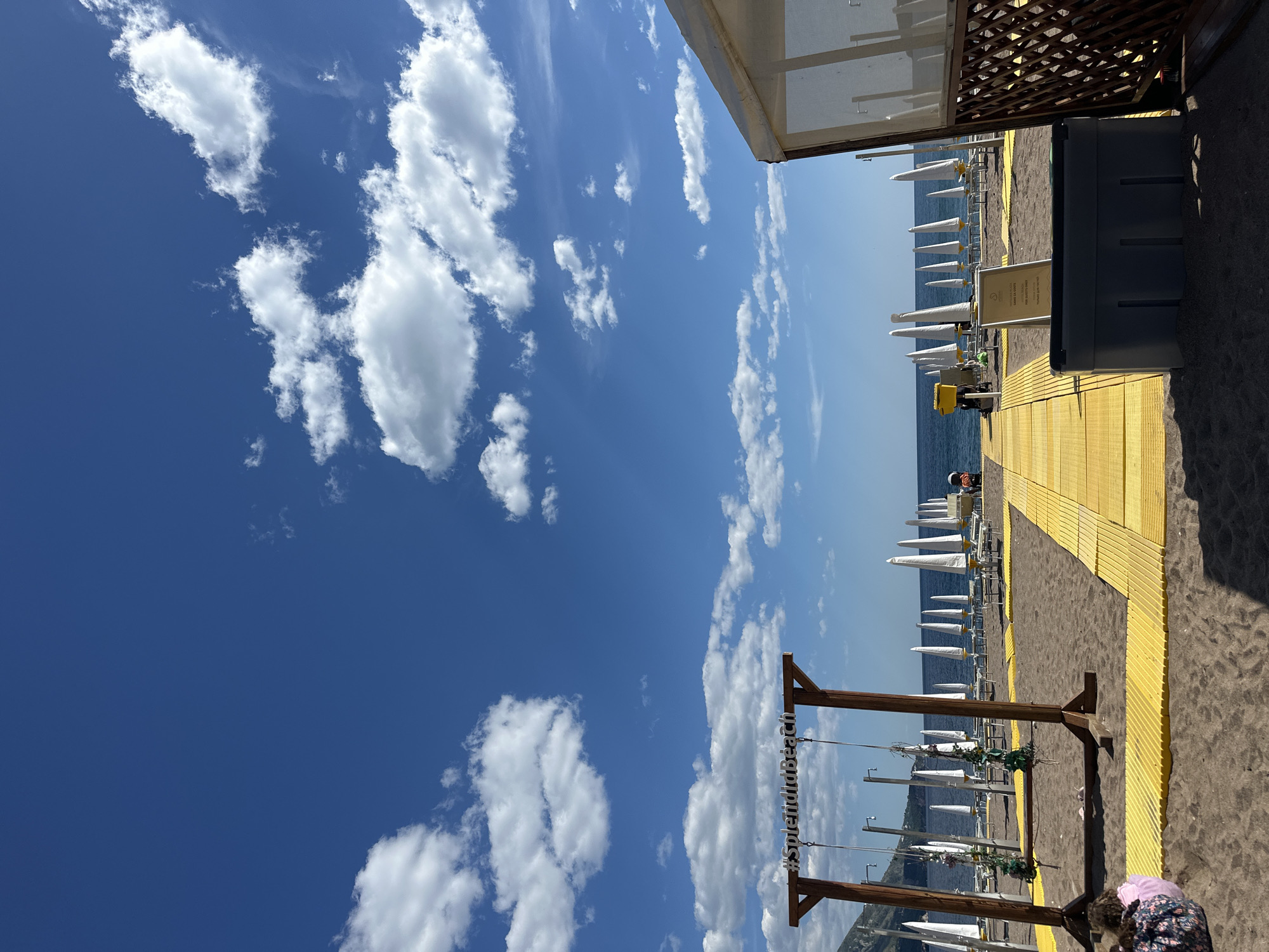 The image depicts a vibrant and sunny beach scene, likely at a resort, under a clear blue sky dotted with scattered white cumulus clouds. The perspective is slightly elevated, looking towards the sea.
In the foreground, the beach features dark sand. A prominent yellow wooden walkway or path extends from the right side of the frame, leading towards the water. To the left of this path, rows of neatly arranged white beach umbrellas and sunbeds line the shore, stretching towards the horizon. The sea in the distance appears calm and blue, meeting the sky at a clear horizon line. Across the water, distant green hills or land masses are visible.
On the right side of the image, parts of a modern beach facility are visible, including a light-colored awning or building structure. A large, dark grey box, possibly a storage unit or waste container, sits on the sand next to the yellow path. Near this box, a sign with text in both English and Cyrillic script is partially visible. The English text seems to indicate rules or information, possibly including "NO ENTRY" or similar warnings, while the Cyrillic text appears to state "ПЛЯЖИ ДЛЯ ГОСТЕЙ" (Beach for Guests).
A rustic wooden frame, resembling a swing set or a decorative arch, stands on the beach near the bottom center. Hanging from it are decorative elements, possibly flowers or greenery. The text "#SplendiduBeach" is clearly visible on a sign attached to the crossbar of this wooden frame.
There are a few people visible. In the bottom right corner, a person, possibly a child, wearing pink and floral attire, is partially obscured. Further down the yellow path, near the water, another individual wearing a white hat can be seen.
The time of day is bright daylight, suggesting mid-morning to early afternoon, with excellent weather for beach activities. The overall atmosphere is one of leisure and relaxation, typical of a well-maintained private or hotel beach. Based on the location context provided, this scene is in Boreti, Montenegro.