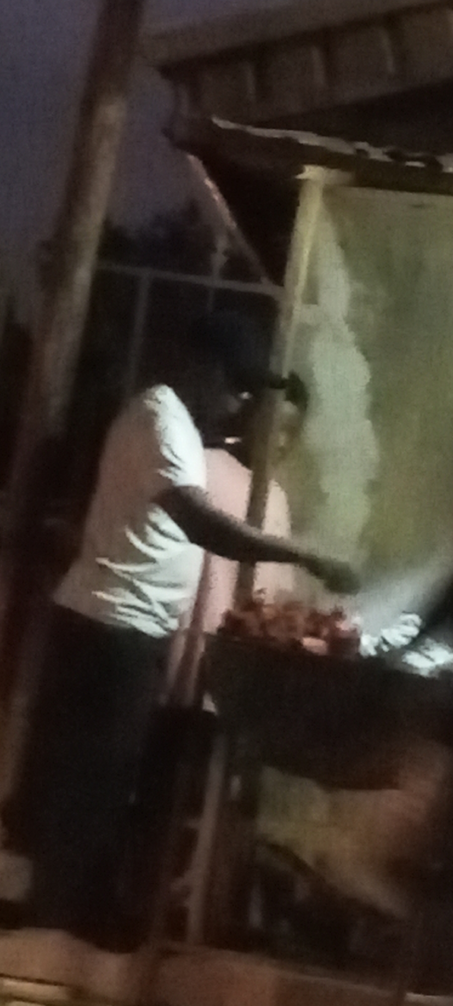 In a dimly lit outdoor setting in Sabon Tasha, Nigeria, two individuals are positioned behind a barbecue grill. One person, wearing a light-colored t-shirt and dark pants, extends their arm over the grill, seemingly tending to the food. Smoke rises from the grill, obscuring some of the contents. The surrounding area includes a makeshift shelter constructed from wooden supports and a light-colored sheet.