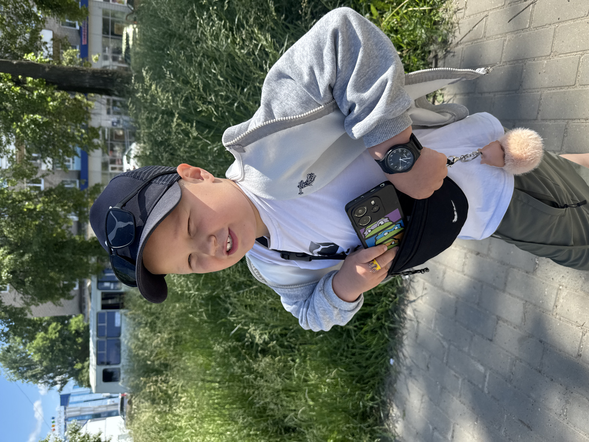 A young boy wearing a black baseball cap and sunglasses stands outside, holding a smartphone and a fluffy keychain. He has a ring on his finger. The boy is wearing a white t-shirt, a grey zippered jacket, and olive-green shorts. He has a black fanny pack around his waist. A black watch is on his left wrist. He is standing on a paved sidewalk, with tall green grass and trees in the background. In the distance, there is a building and what appears to be a tram. The scene is lit by sunlight, casting shadows on the sidewalk. The location is the city of Chaikovsky, Russia.