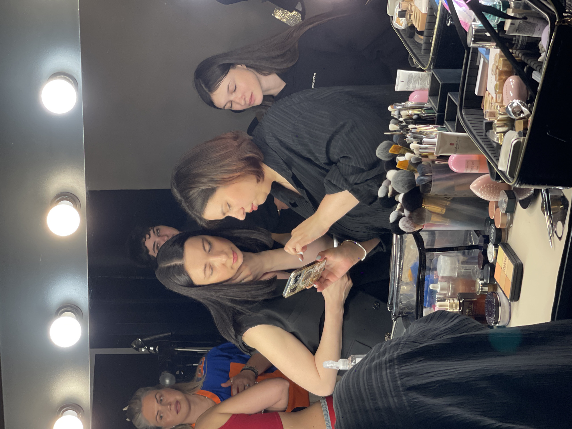 In Kyiv, Ukraine, a group of women are gathered around a vanity mirror illuminated by bright bulbs. Two women in the foreground are focused on a smartphone held by the woman on the left, who is looking down at the screen. The woman on the right, with her eyes closed, is positioned behind a collection of makeup brushes and cosmetic products laid out on the vanity. To their left, another woman, wearing an orange and blue top, looks towards the mirror. Behind them, a fourth woman with dark hair is partially visible. The vanity is laden with various makeup items, including brushes, palettes, bottles, and containers, suggesting a professional makeup application or preparation setting. The overall environment appears to be an indoor space with dark walls.