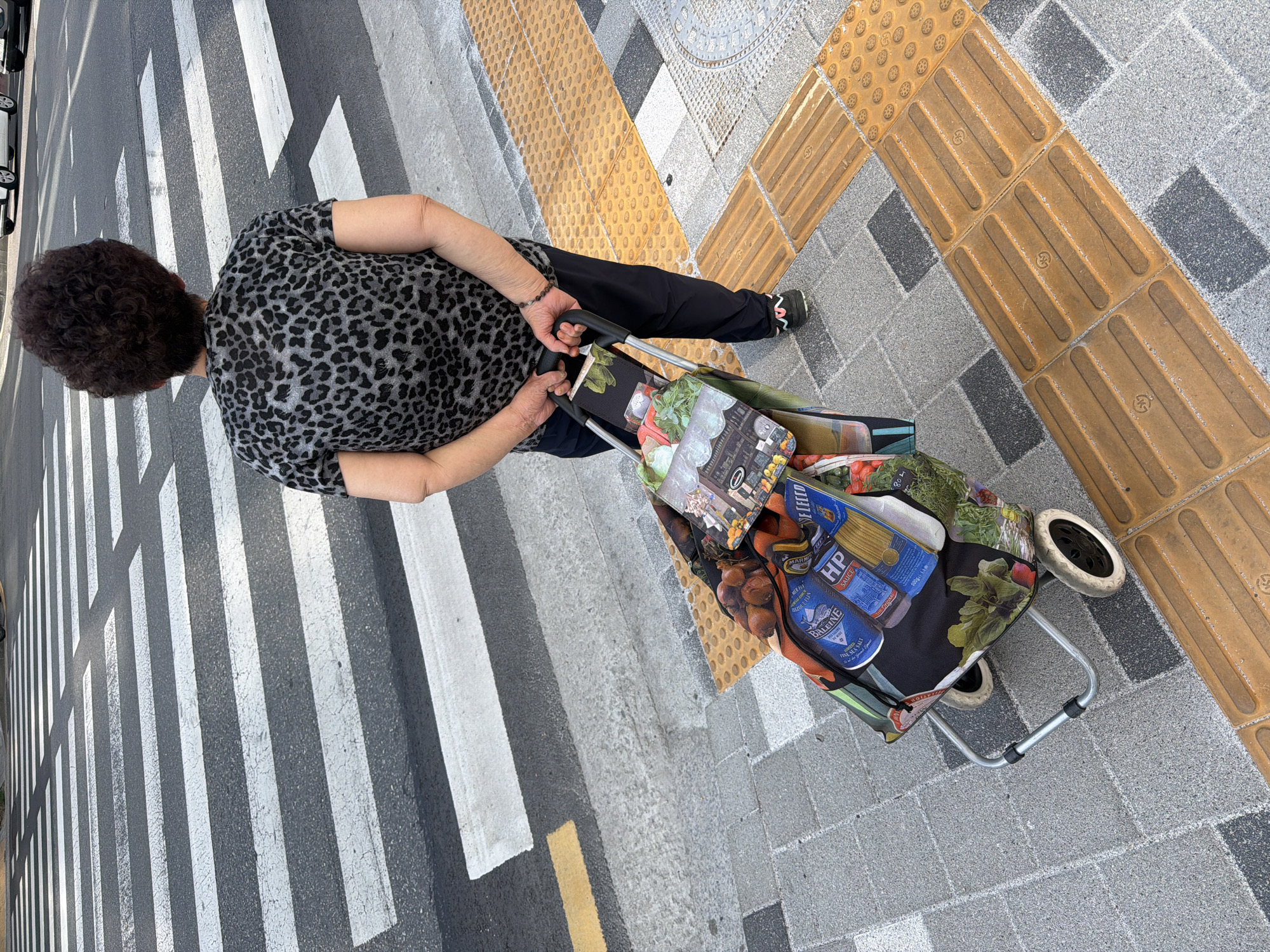 A woman stands on a crosswalk and pulls a two-wheeled shopping cart on a street in Seoul, South Korea. The crosswalk has alternating gray and white stripes, with a yellow painted line on one side. The woman has dark, curly hair and wears a gray and black leopard-print top and black pants. She has a bracelet on one wrist. She pulls a shopping cart that has groceries inside. The shopping cart has a colorful bag that has grocery store packaging printed on it. The cart has two wheels. The ground is gray pavement. There are raised yellow pavers adjacent to the sidewalk.