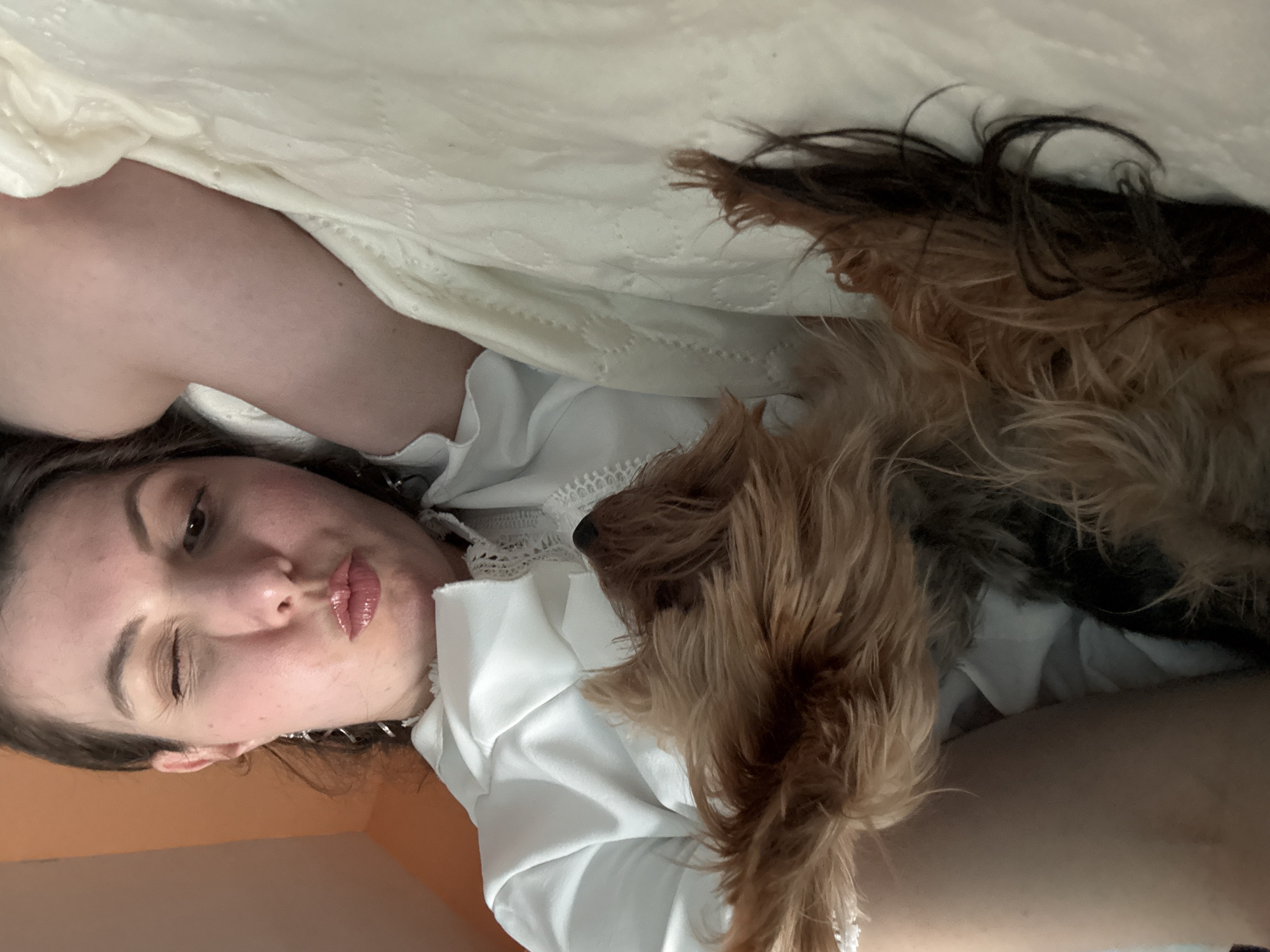 A woman lies sideways in a bed with a small dog. The woman is facing the camera with pursed lips and one eye closed. She has brown hair and is wearing a white top with lace detailing. A cream-colored blanket is pulled up towards her shoulder. The dog, brown and black, lies near her chest, its head facing to the right. A tan-colored wall is visible in the background. The location is Unknown, Unknown.