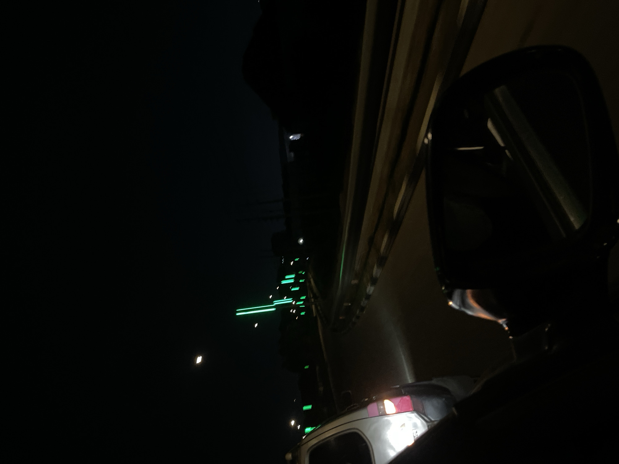 A nighttime image taken from inside a vehicle. The vehicle's side mirror is visible in the right part of the frame, reflecting the dark environment. A vehicle's rear lights appear at the lower center. In the left background, there are illuminated green bars on a tower, appearing to be telecommunications. The moon is barely visible in the left side of the image.
Given the location is Jiru, Nigeria, the tower could be a cell phone tower or other broadcast tower.