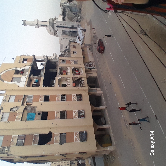 The image depicts a devastated urban street scene in Khan Yunis, Palestinian Territory, captured from an elevated perspective during daytime. The central focus is a multi-story residential building on the left, which has suffered extensive damage. Its facade is riddled with holes, many windows are broken or missing, and sections are completely gutted, exposing interiors filled with debris. Other damaged buildings are visible further down the street, and in the background, a white mosque with a prominent minaret also shows signs of damage or disrepair, surrounded by rubble.
A paved road, marked with white lines, runs through the scene. A red car is parked on the road, and a small group of approximately five individuals, including men and possibly boys, are seen walking or standing on the street. Their attire is casual. One person, partially visible in the top right, appears to be taking the photo. The sky is bright, suggesting daytime conditions. The overall impression is one of severe destruction in an urban environment. The text "Galaxy A14" is visible in the bottom right corner of the image, identifying the camera device.