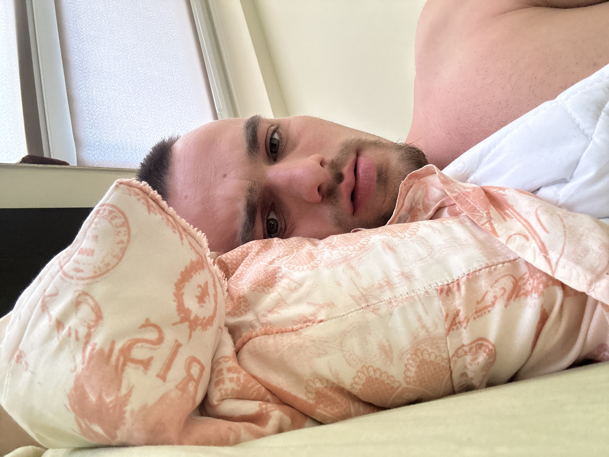 A close-up, low-angle shot depicts a man reclining in bed. He is fair-skinned with dark hair and stubble. He is partially covered by a pink and white patterned pillow and a white blanket or sheet. His bare shoulder is visible. A window with blinds or frosted glass is in the background. The exact location in Sofia, Bulgaria cannot be determined from the image.