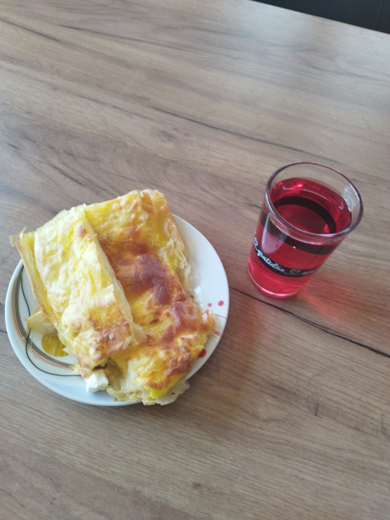 On a wooden surface in an unknown location, there is a plate of food and a glass of red liquid. The plate, primarily white with patterned brown and yellow accents, holds a pastry that appears to be a cheese burek or similar baked dish. The pastry has a golden-brown exterior and seems to be layered with cheese filling. Adjacent to the plate is a clear glass filled with a red liquid, possibly juice or a soft drink. The glass has a white design on its side. The wooden surface provides a neutral backdrop to the food and drink.