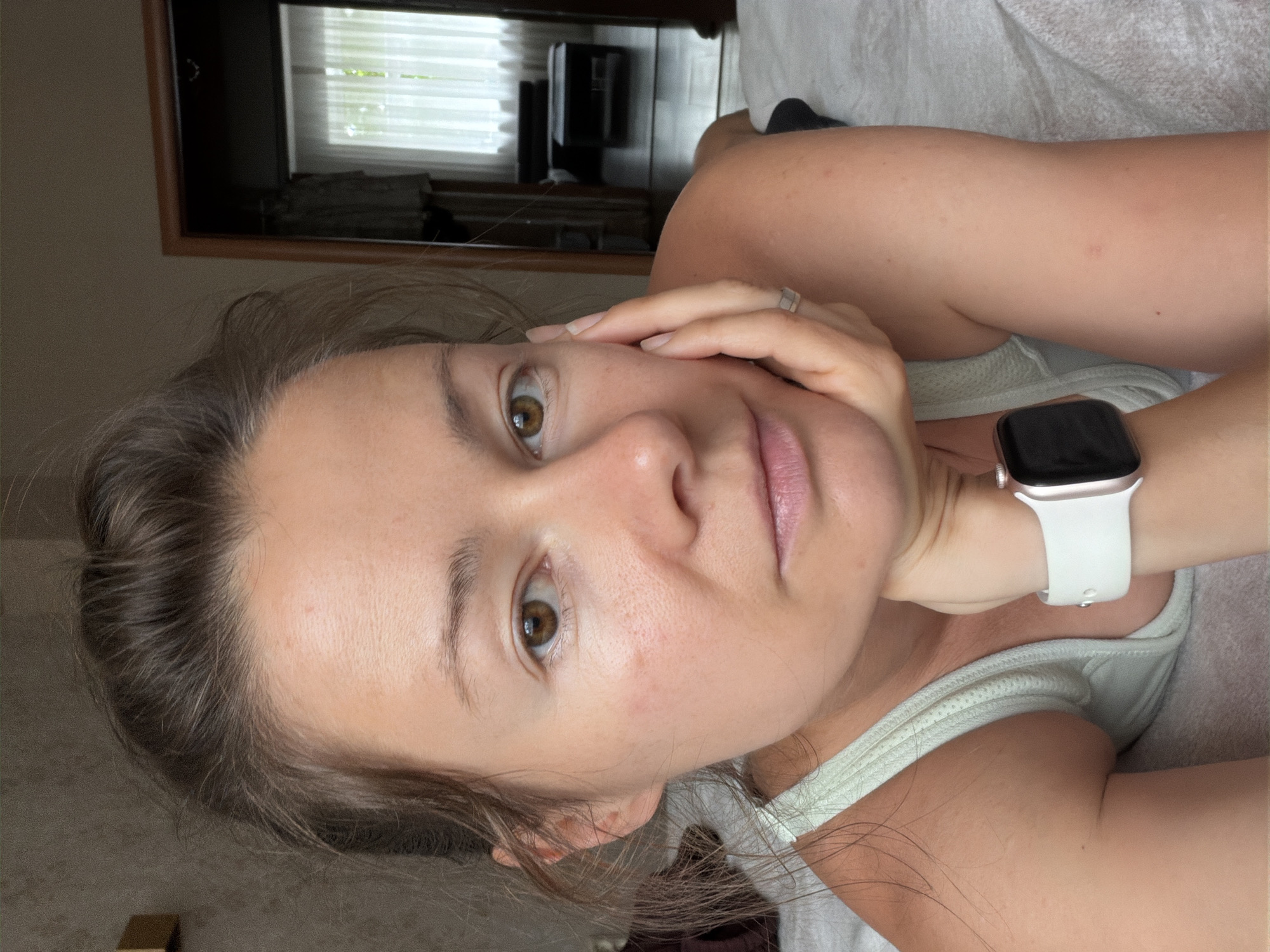 A woman with brown hair tied back is lying on a bed, resting her head on her hand. She is wearing a light green tank top and a white smartwatch. The room appears to be a bedroom with a doorway in the background leading to another room with a window and furniture. The overall impression is of a casual, indoor setting. The image does not provide any information about the city of Riga, Latvia.