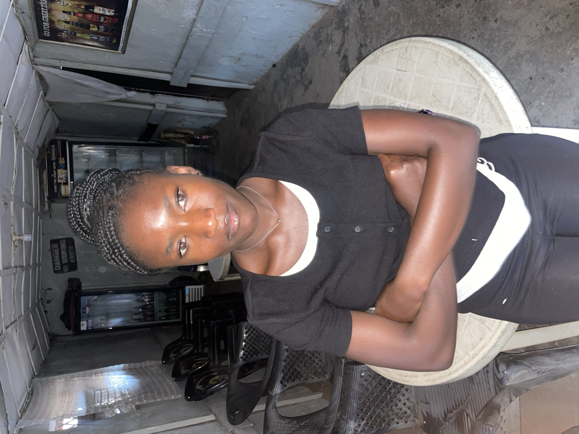 The image features a young, dark-skinned woman seated indoors, likely in a casual bar or eatery in Jos, Nigeria. She has braided hair (cornrows) and gazes directly at the camera with a neutral expression. She is dressed in a black, short-sleeved button-up top layered over a white undershirt, complemented by black pants or leggings with a distinctive white stripe on the side. A thin silver necklace is visible around her neck, and her arms are crossed.
She is positioned on a light-colored, possibly plastic, stool or low table with a textured, grid-like surface. The background reveals a somewhat dimly lit interior space. Notable objects include several dark-colored chairs, some appearing stacked, and a glass-fronted refrigerator or display cooler containing various bottles, presumably beverages. The walls are constructed from light-colored, painted blocks, and the floor is dark and uneven, featuring a metallic grate. A piece of white fabric hangs from the ceiling structure.
The artificial and dim lighting suggests the photograph was taken in the evening or at night. The woman is the central subject, calmly posing for the camera within this local setting.
Several textual elements are visible in the background:
- A poster on the upper wall partially reads "GO FOR GREAT TAS..." (likely "TASTE") and displays images of assorted beverage bottles.
- A smaller poster near the refrigerator clearly states "START THE NIGHT WITH GUINNESS".
- A black sign mounted on the wall explicitly warns "DANGER NO SMOKING AREA".