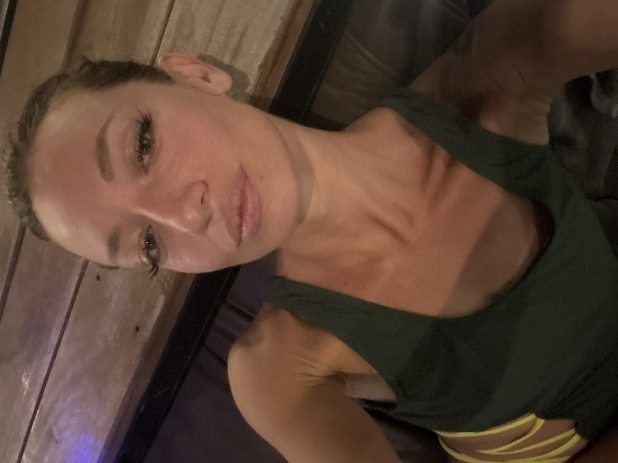 A selfie-style medium close-up shot features a young adult female, captured from the chest up with her head tilted towards the upper left of the frame. She looks directly at the camera with a slight, relaxed smile. Her hair is styled back, and she displays prominent eyelashes and well-defined eyebrows.
She is wearing a dark green tank top or swimsuit top. Below the green top, the yellow, multi-strap design of a bikini bottom is visible. The woman has a fair complexion, with noticeable redness or tan lines across her upper chest and shoulders.
The lighting in the image is dim and warm, creating soft shadows and highlights. The background to her left and above her head consists of vertical wooden paneling, indicating an indoor or covered outdoor setting. Dark, indistinct areas fill the rest of the background, with a faint blue light emanating from the bottom left corner. The scene suggests a relaxed atmosphere, potentially within a resort or private dwelling, fitting the context of บ้านบางรักษ์, Thailand. There is no visible text in the image.