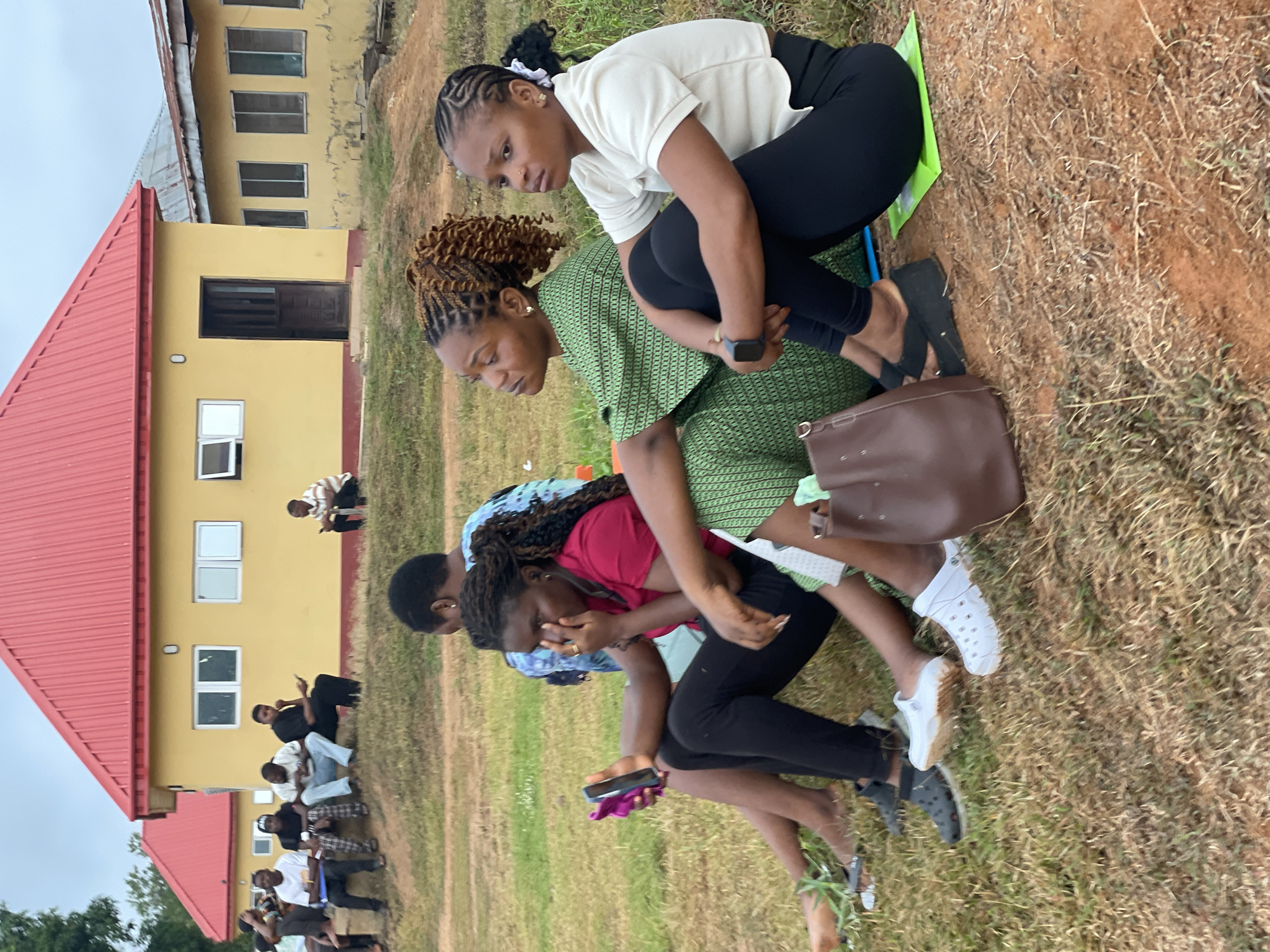 The image shows a group of people sitting on the ground outside a yellow building with a red roof. Several people are in a "piggyback" like position. The location is Ise - Ekiti, Nigeria. The environment appears to be a grassy area or field. Several people are sitting on the ground near the buildings. The group of people in the foreground are sitting very close together.