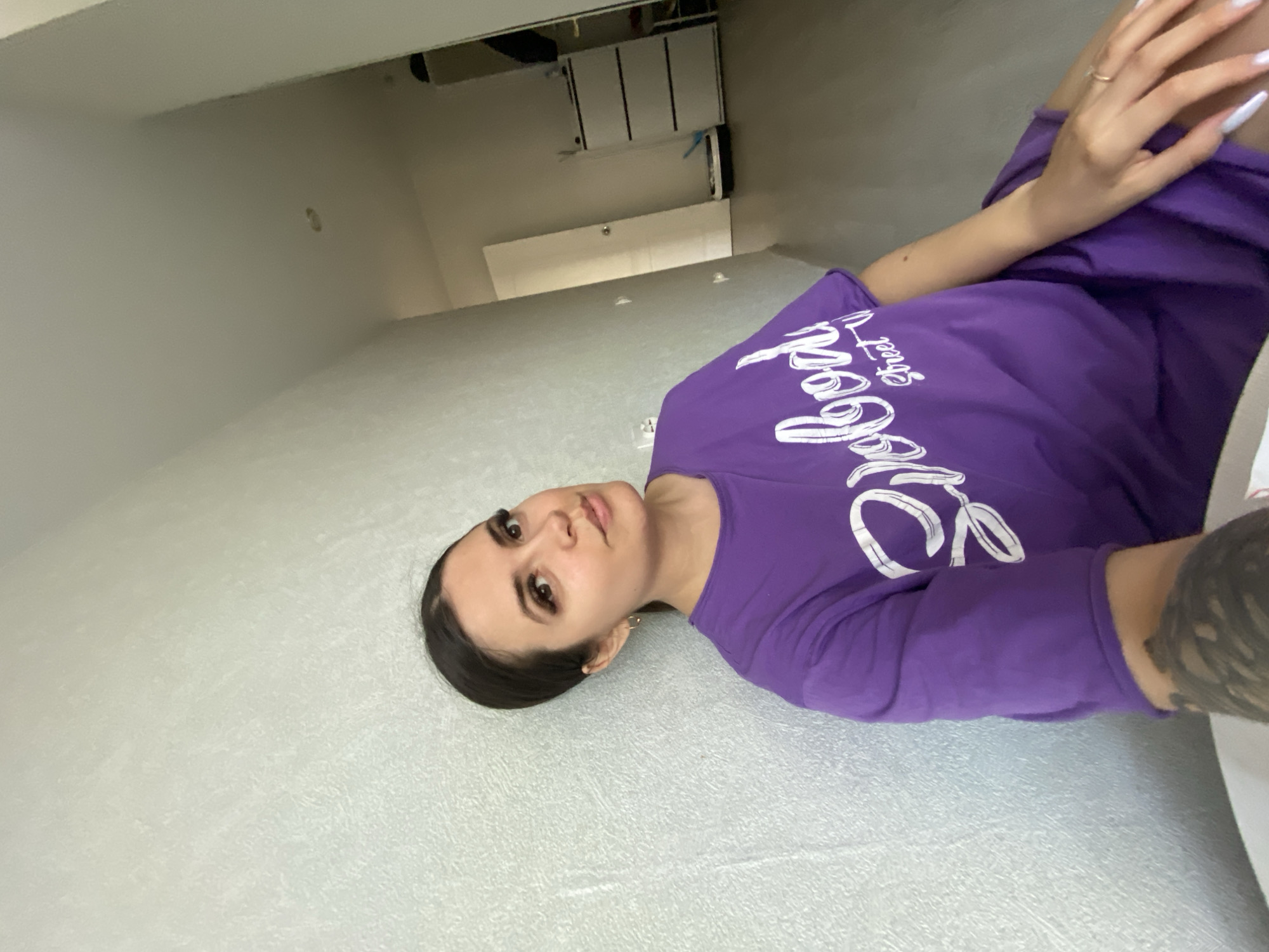 The image shows a woman with dark hair and brown eyes wearing a purple long-sleeved shirt with white writing. She is lying on a grey textured surface, possibly a bed or carpet. Her left arm is visible, featuring a tattoo. The background indicates an interior space with white walls, a door, and some cabinets. The location is in Chaikovsky, Russia.