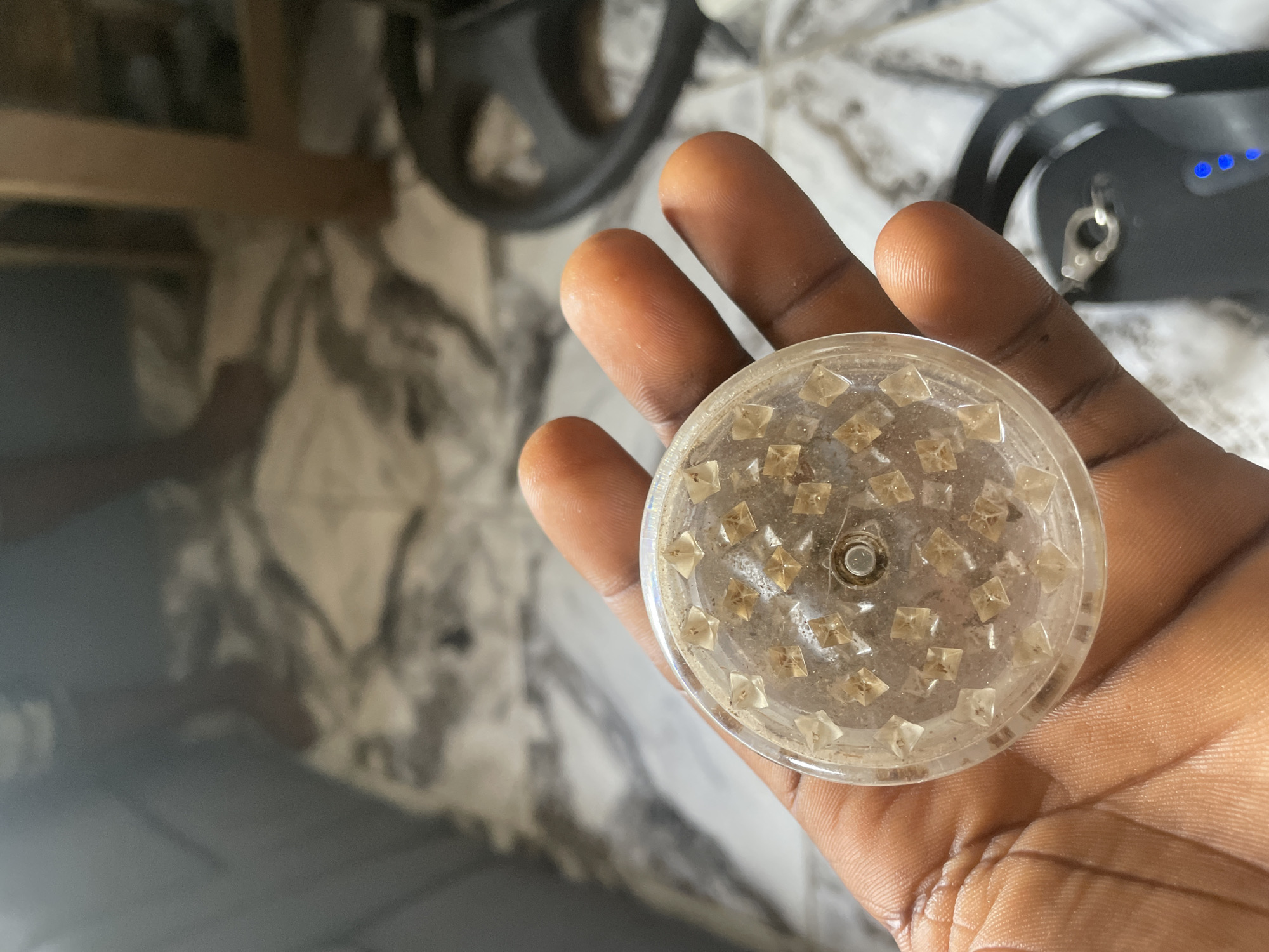 A close-up, eye-level shot shows a person's dark-skinned hand holding a clear, circular object with pyramid-shaped teeth. The object appears to be a grinder, possibly for herbs or spices, and contains some residue. The background is blurred, but suggests an indoor environment with a tiled floor and furniture, consistent with a residential setting in Keffi, Nigeria. The lighting is dim, creating a slightly grainy effect.