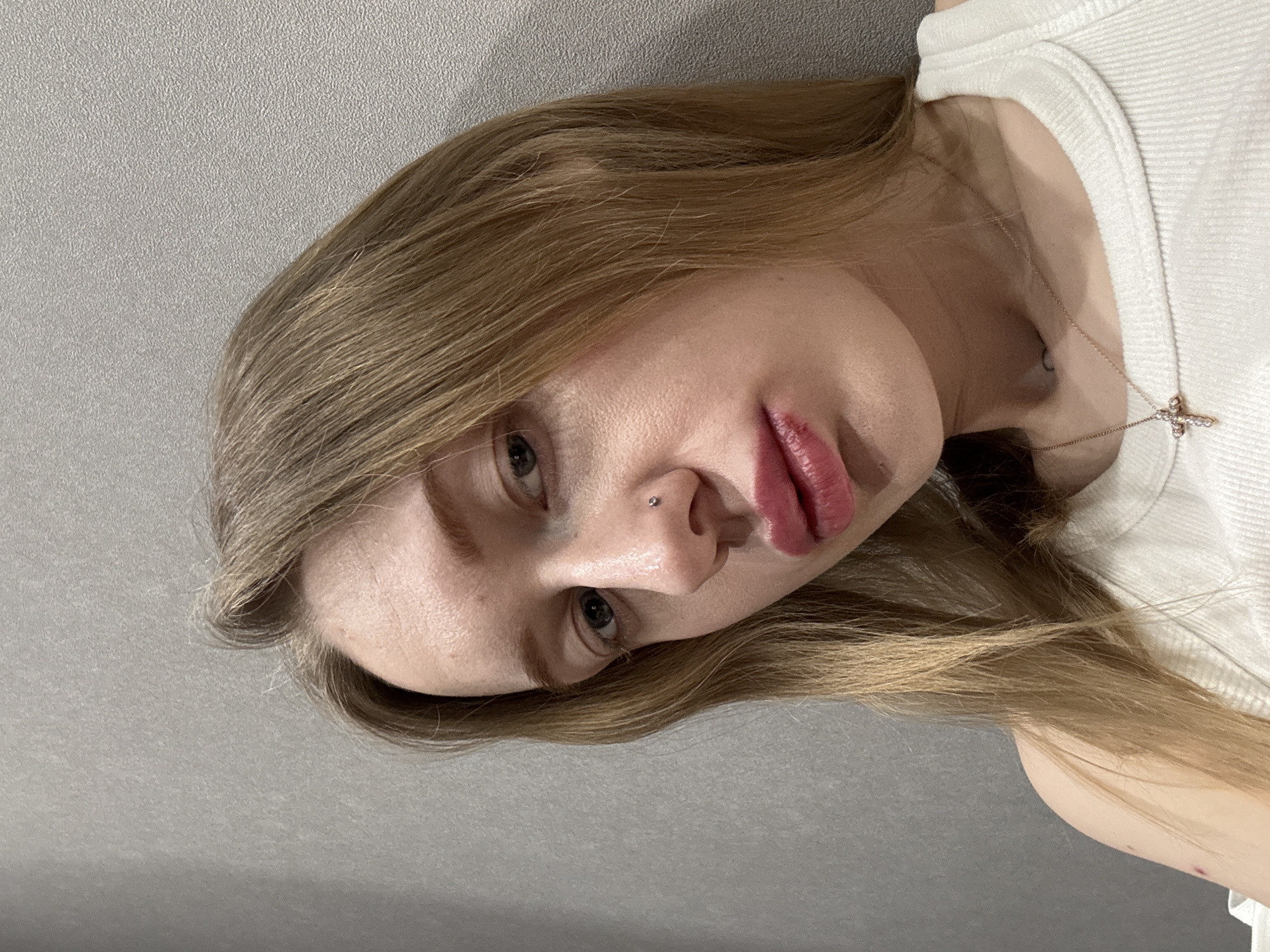 A close-up, head-and-shoulders portrait of a fair-skinned female subject is depicted against a plain, light grey, textured wall. The location is stated as Rostov-on-Don, Russia. The subject, with long, straight, light brown hair parted down the middle, looks directly into the camera with blue-grey eyes. Her nose features a small silver stud piercing on the left nostril. Her lips are full and colored with red/pink lipstick, with a visible irregularity or bruise on the left side of the upper lip. She wears a white ribbed tank top. A thin, gold-colored chain necklace with a small, ornate, jewel-encrusted cross pendant rests on her sternum. A small, dark, circular tattoo is visible on her upper chest, partially obscured by the necklace. A small red mark or blemish is present on her exposed left shoulder/upper arm. The lighting is uniform, suggesting an indoor environment. No other subjects or objects are visible.
